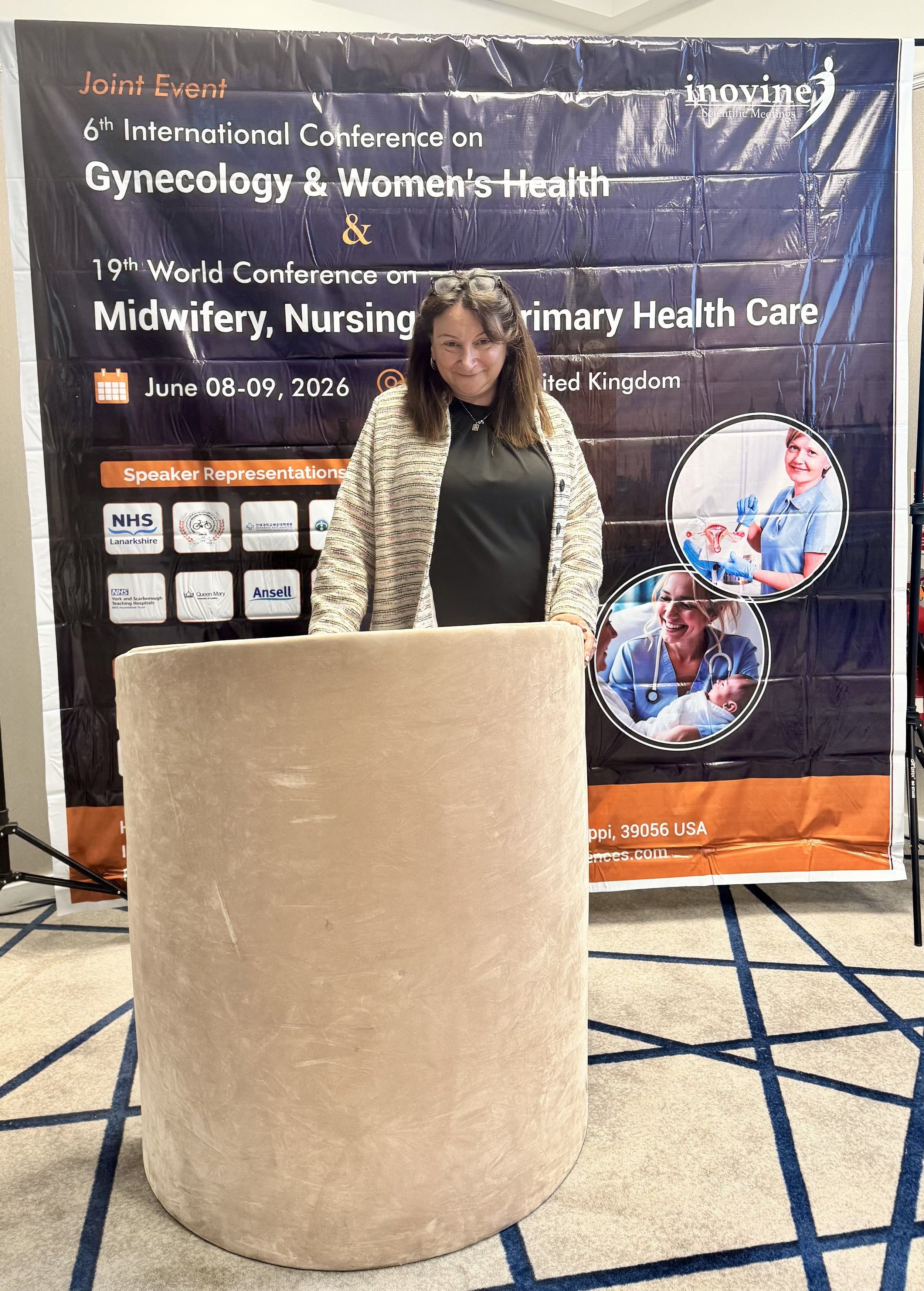 A woman standing in front of a conference banner at a medical event. The banner reads 'Joint Event 6th International Conference on Gynecology & Women's Health and 19th World Conference on Midwifery, Nursing & Primary Health Care, June 08-09, 2026, United Kingdom' and displays logos of various organizations. The woman is smiling, wearing a black top and a beige cardigan, with glasses on her head.