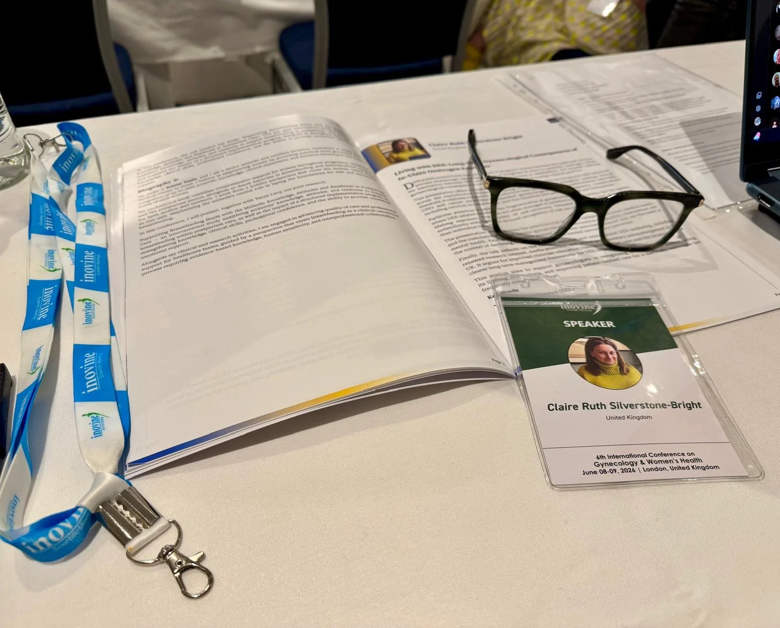 Conference badge for Claire Ruth Silverstone-Bright with her photo and details, glasses, open report or book, lanyard with blue and white logos, and a plastic sleeve with the word "SPEAKER" on it.