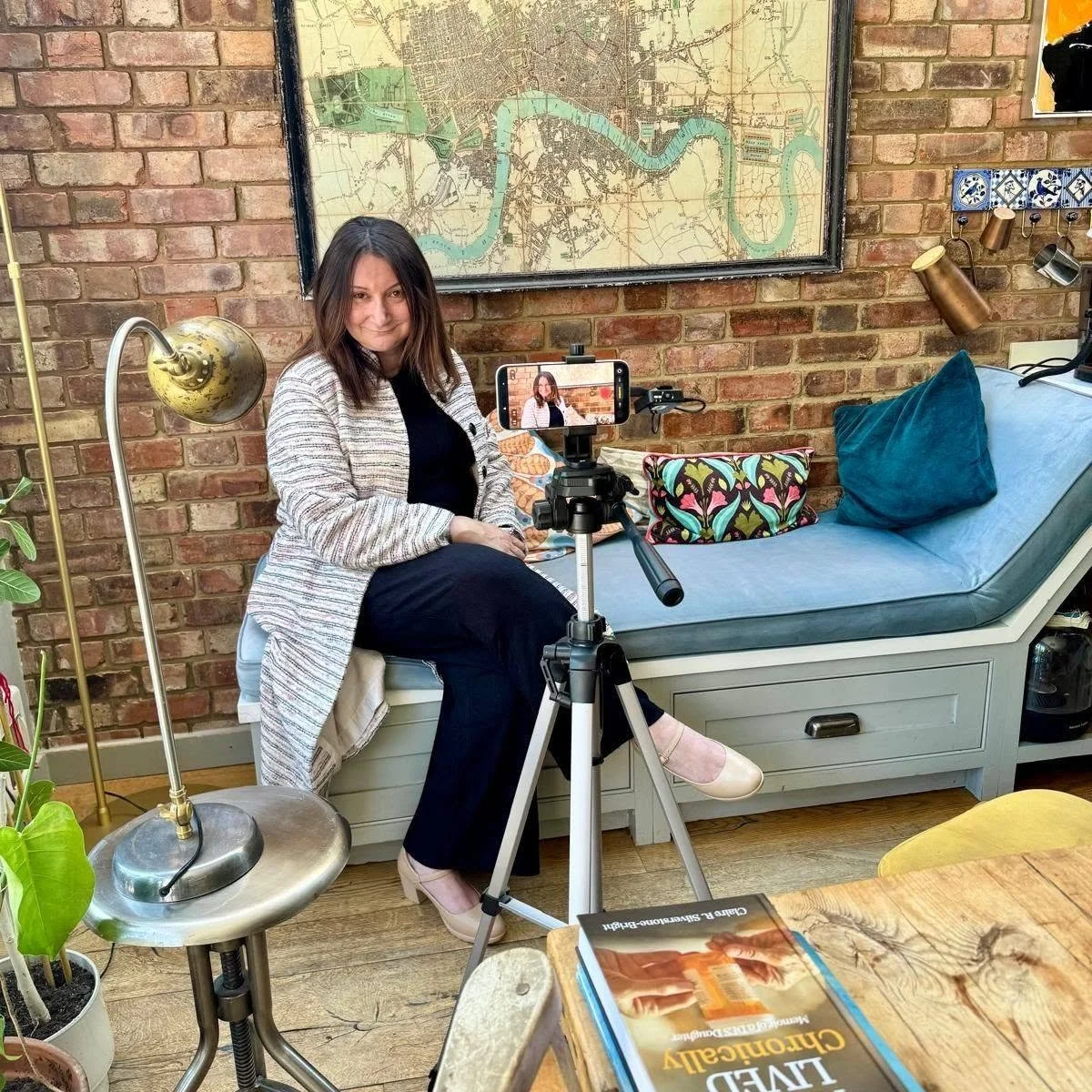 A woman sitting on a bench with 3 colorful pillows, being filmed with a smartphone on a tripod, in a cozy room with a brick wall and a large map behind her, nearby there's a brass lamp, a potted plant, and a wooden table with books.