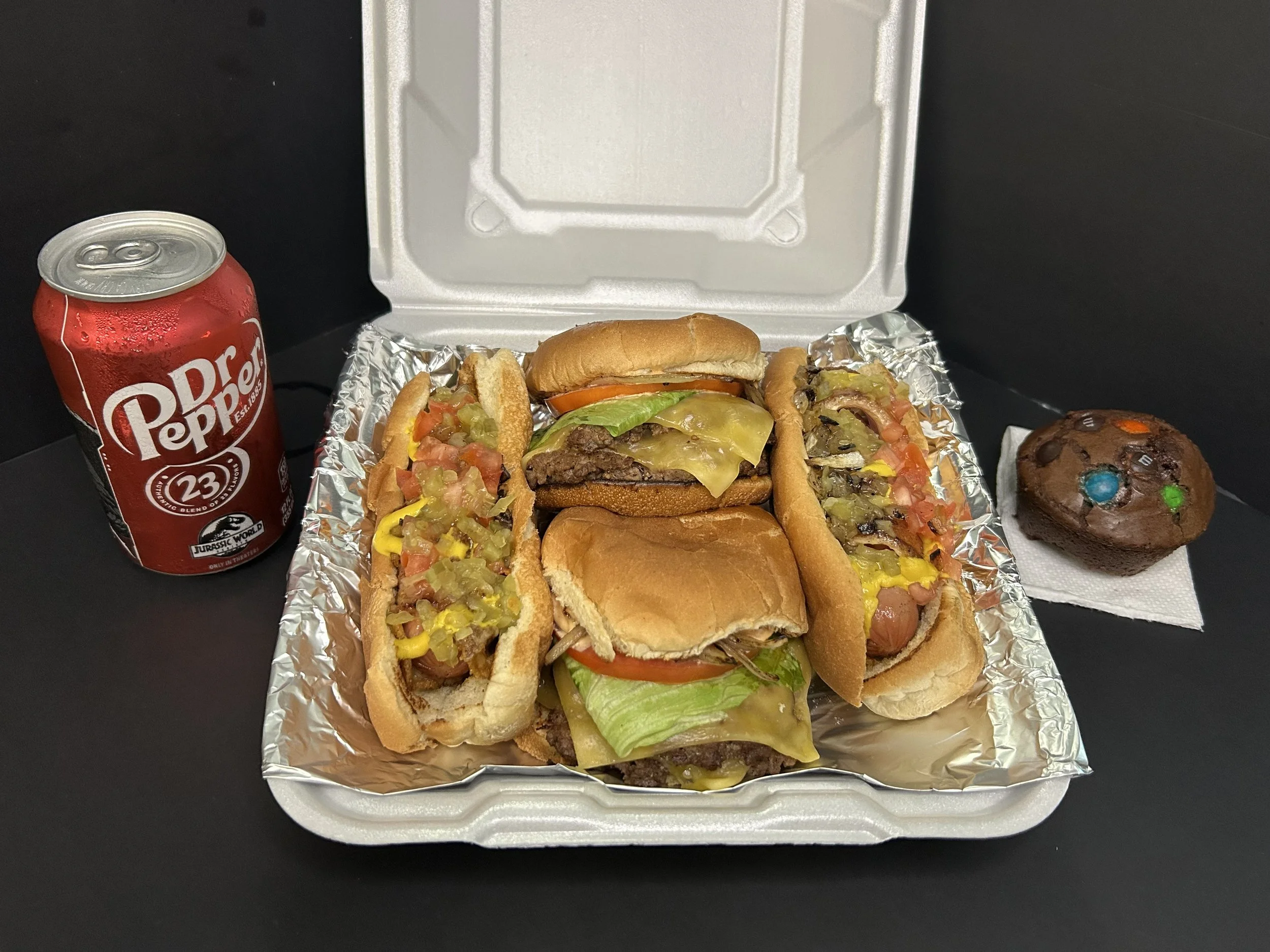 A takeout meal with two cheeseburgers topped with lettuce, tomato, pickles, and cheese, served in bread rolls, with two hot dogs topped with mustard, relish, and chopped onions, on a foil-lined tray. A can of Dr. Pepper soda and a chocolate cupcake with colorful candies on top are also included.