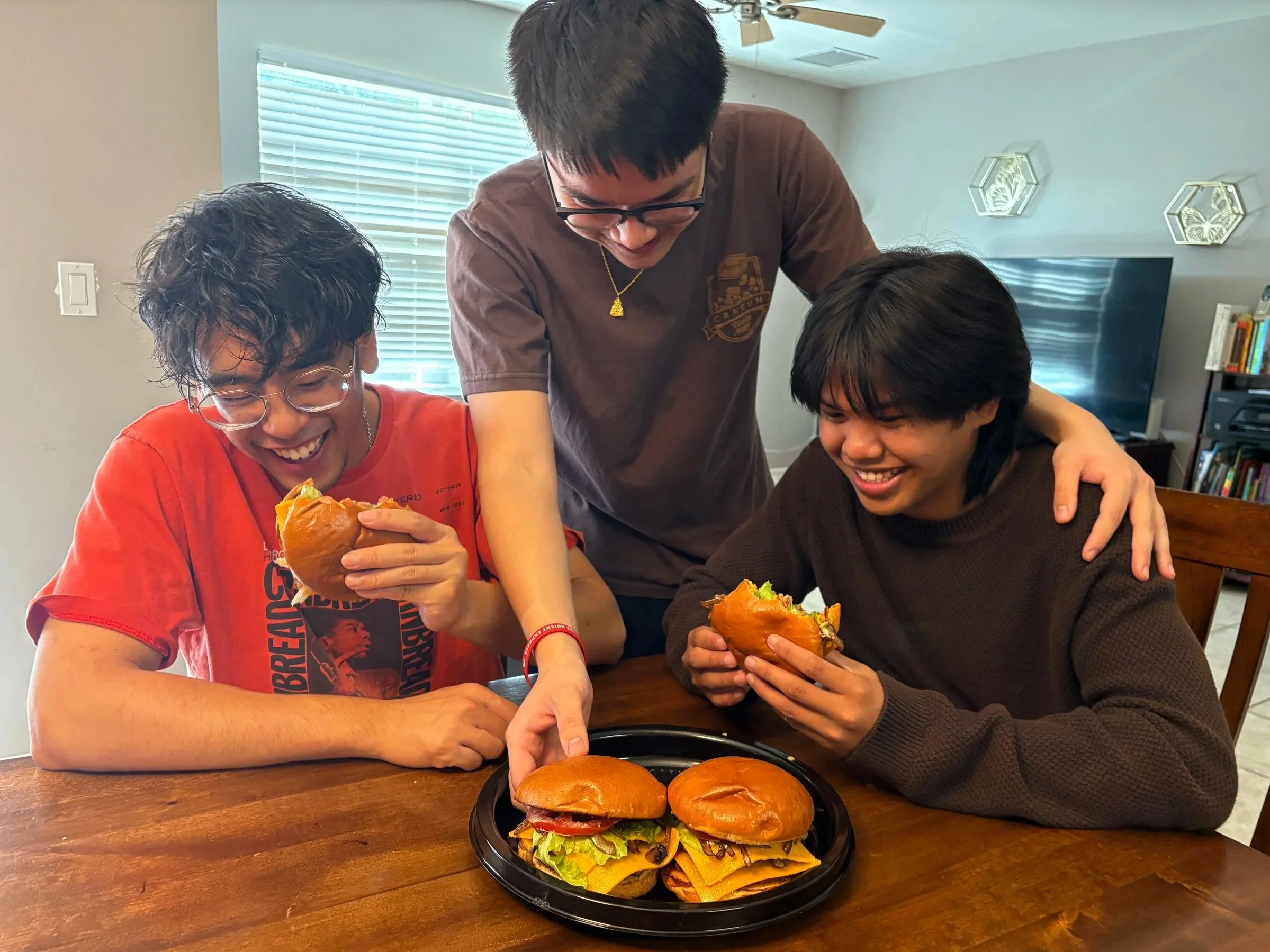 Two young men are sitting at a wooden table, each holding a hamburger, smiling and enjoying their meal with a friend standing between them. There is a plate of two cheeseburgers on the table in front of them. The room has a window with blinds and a TV on a bookshelf in the background.