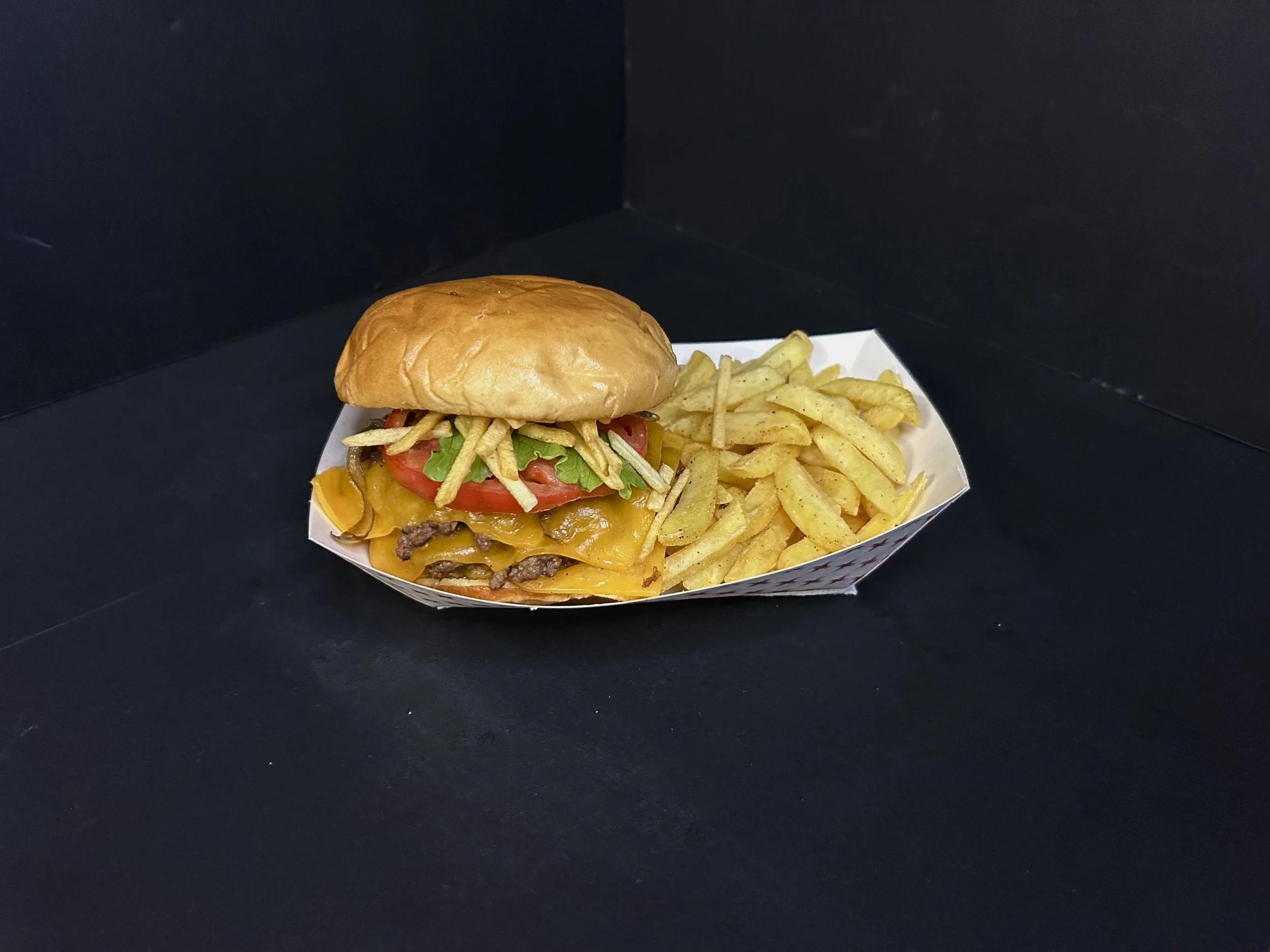 A cheeseburger with lettuce, tomato, pickles, onions, cheddar cheese, and beef patty, served with French fries on a white paper tray.