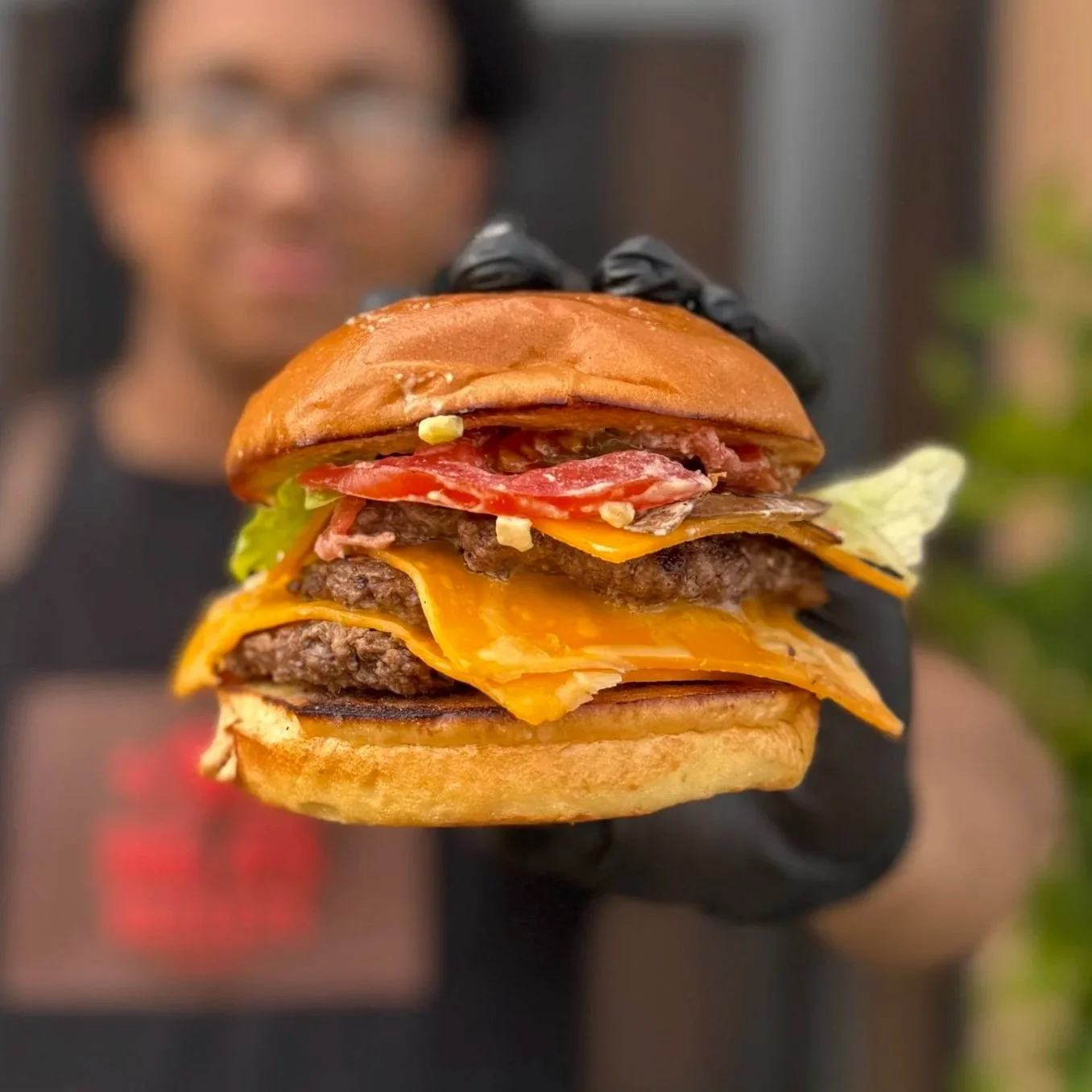 Close-up of a person in the background holding a large cheeseburger with lettuce, tomato, bacon, cheddar cheese, and beef patties, with the person's face blurred.