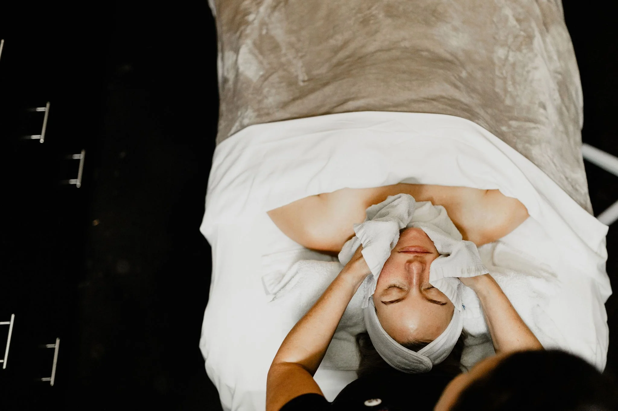 A woman lying on a hospital bed with a towel wrapped around her head, receiving facial treatment with a cloth. She is covered with a white blanket, and a person at the foot of the bed is gently touching her face.