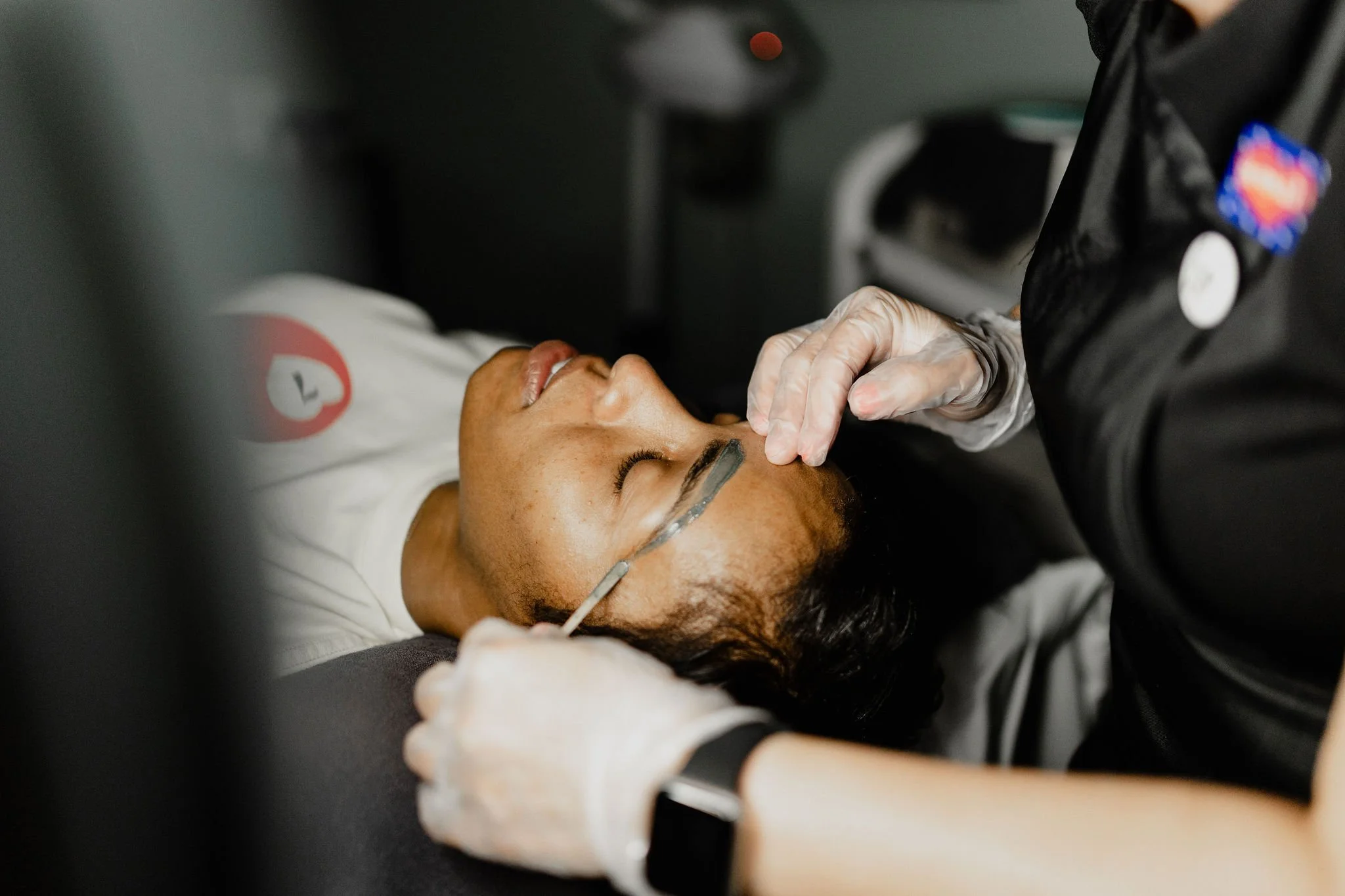 A person lying down with their eyes closed, while a procedure is being performed on their forehead with a small tool by a professional wearing gloves.