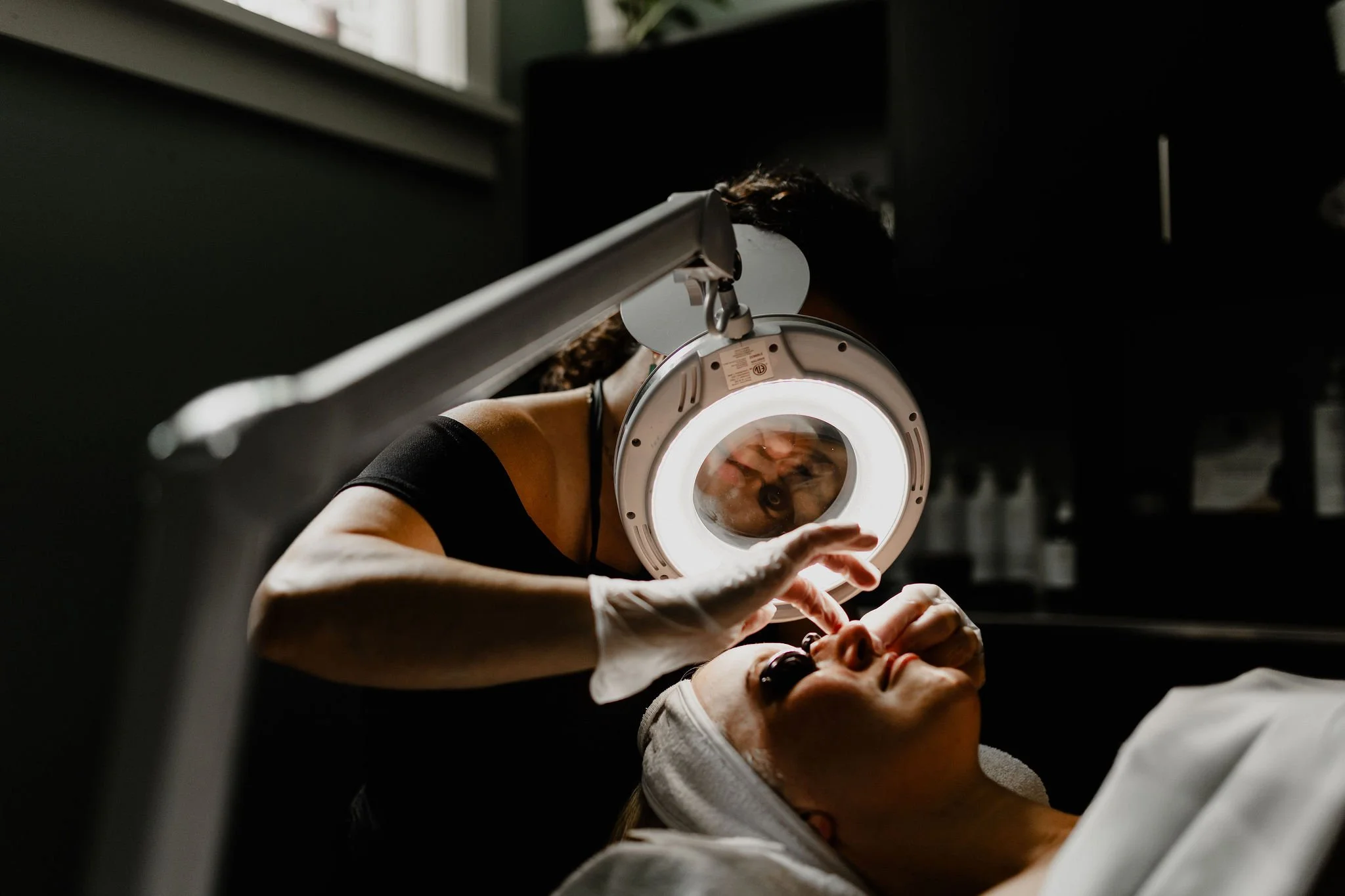 A woman receiving a facial treatment under a magnifying lamp in a clinical or spa setting.
