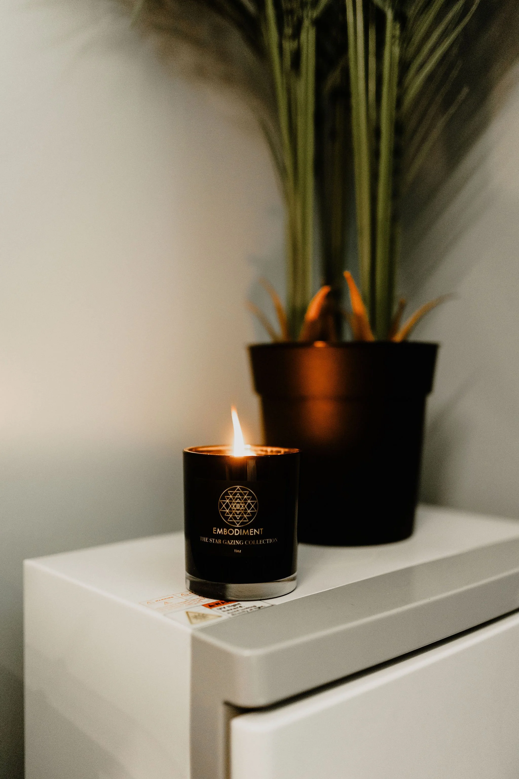 A lit black candle on a white surface with a potted plant in the background.