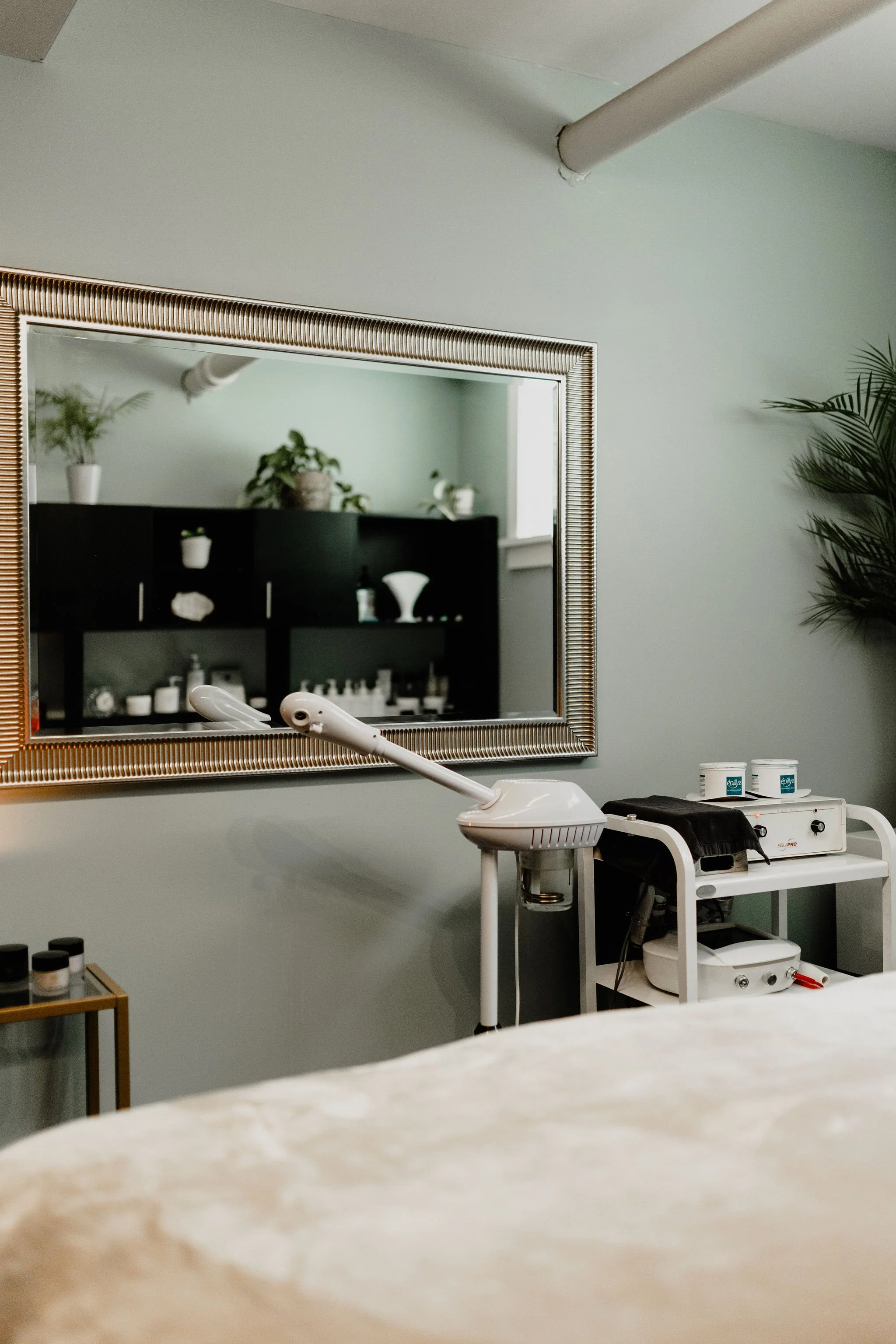 A treatment room with a large mirror, a facial steamer, and skincare products on a cart.