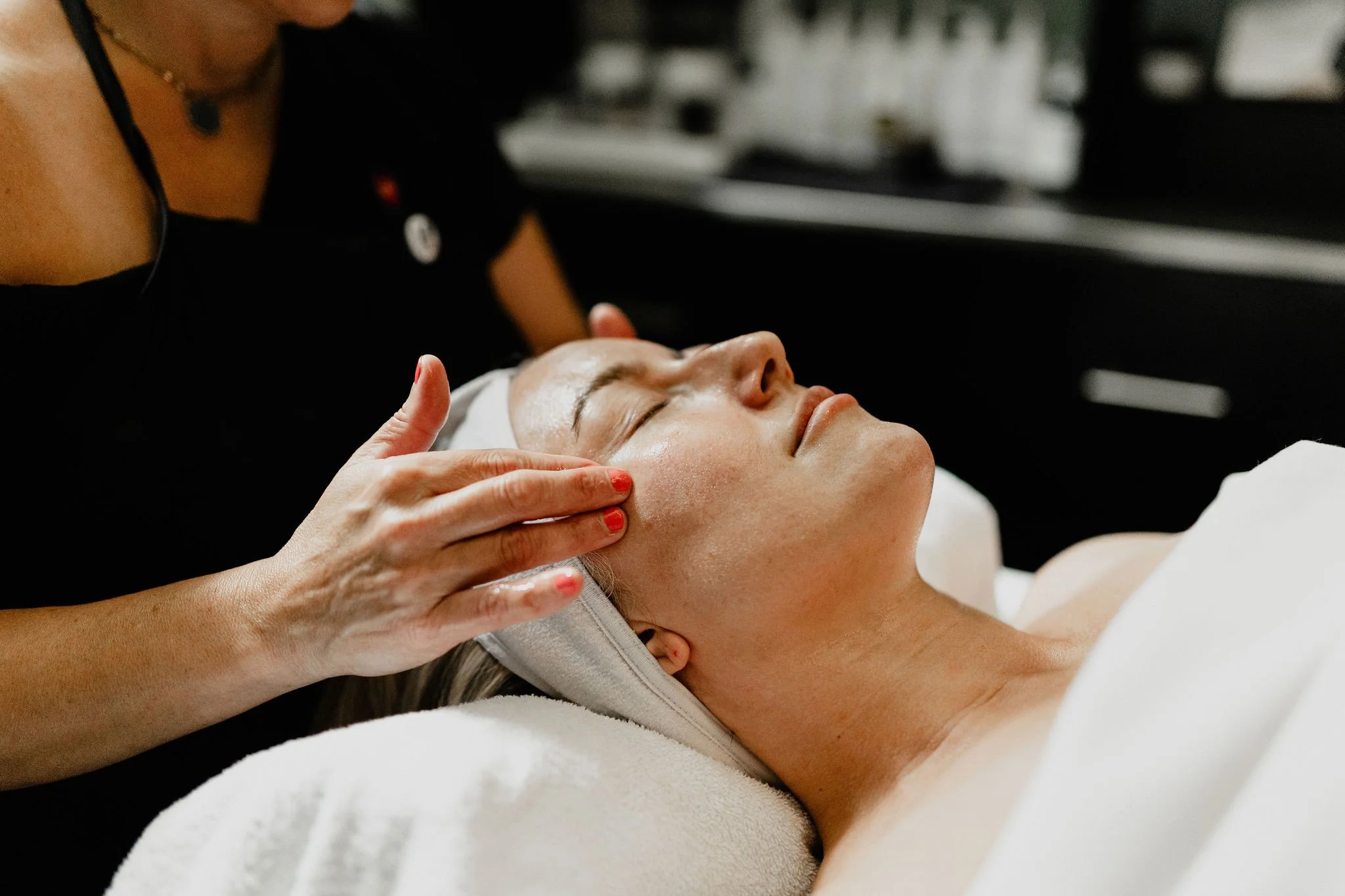 A woman receiving a facial treatment at a spa, lying on her back with eyes closed while a practitioner gently massages her face.