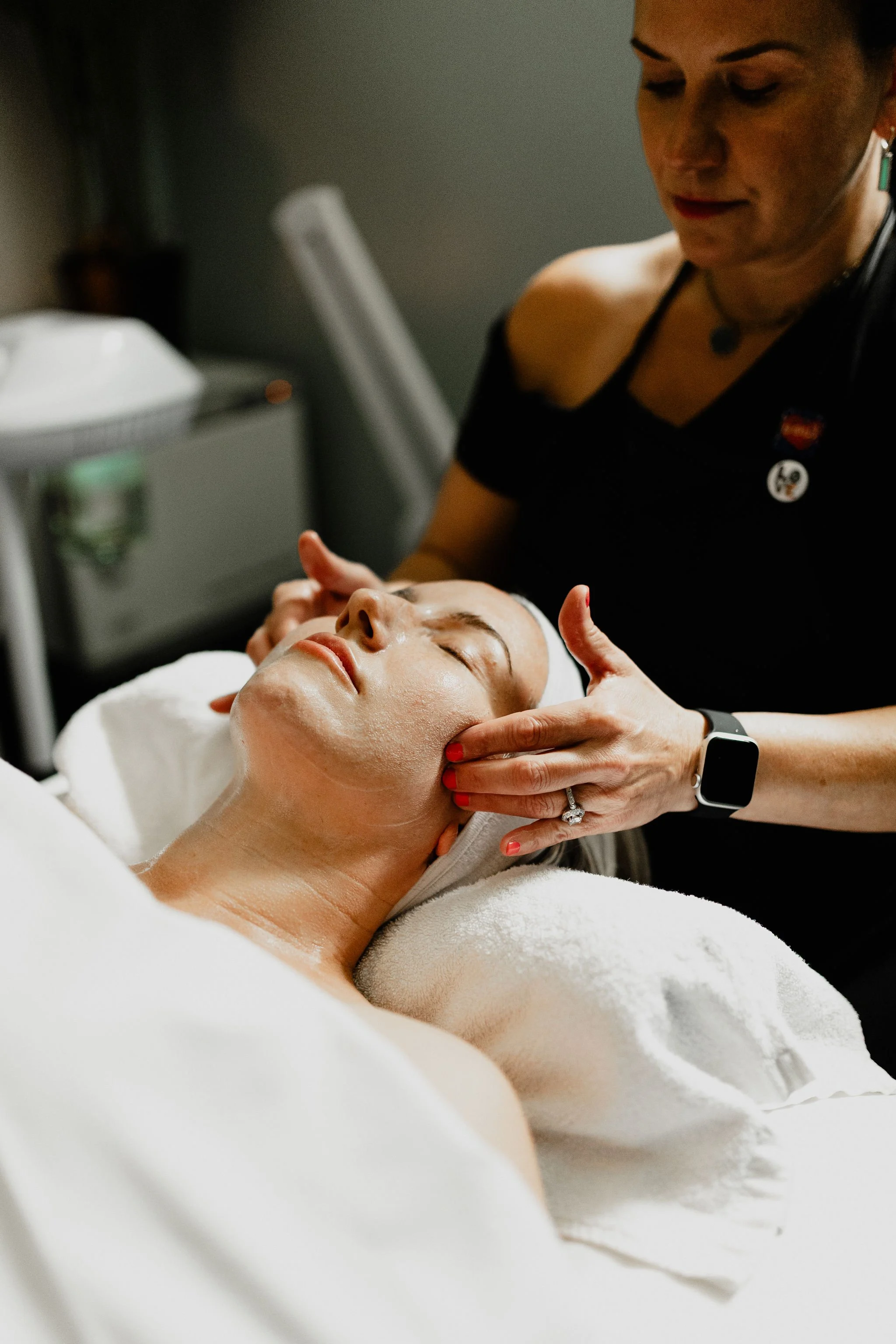 Woman receiving facial treatment at spa, lying down with eyes closed, esthetician gently massaging her face.