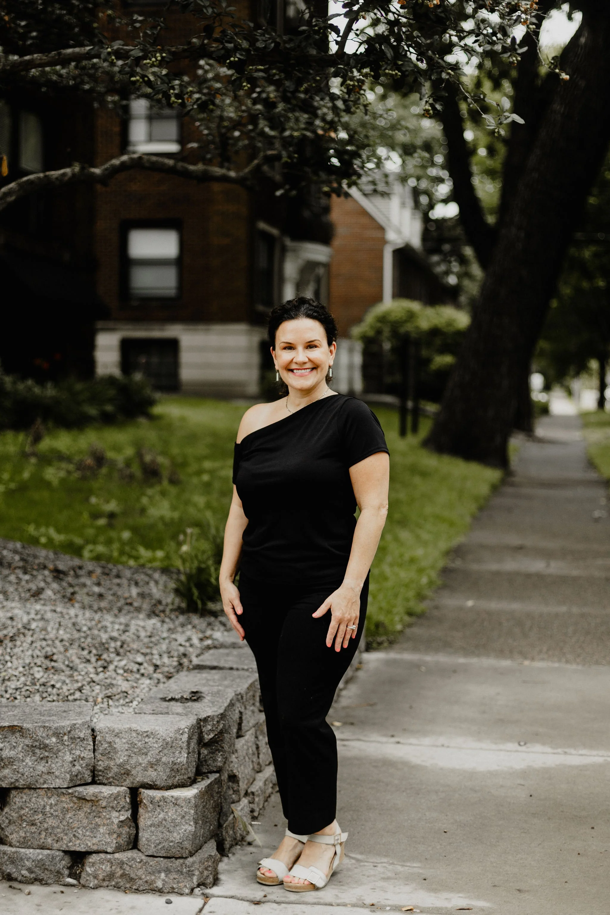A woman in black clothing standing outdoors on a sidewalk next to a low stone wall, with a building and trees in the background, smiling at the camera.