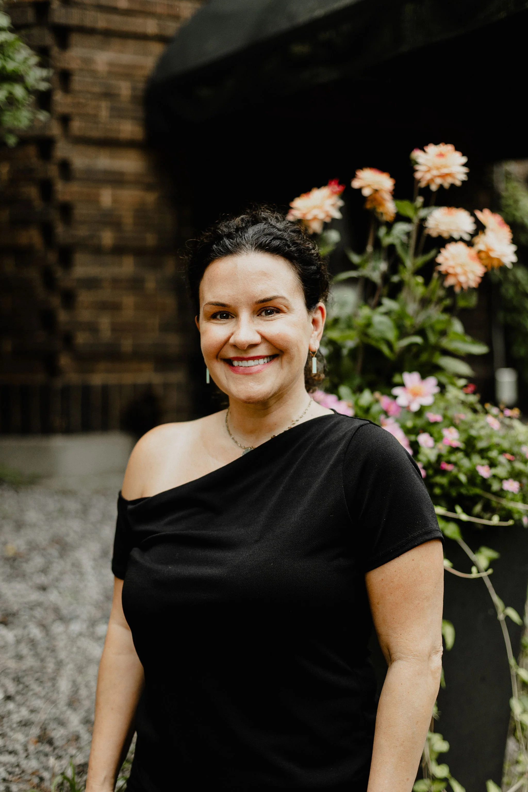 A woman with dark curly hair and a smile, wearing an off-the-shoulder black top, standing outdoors near flowers and a brick wall.