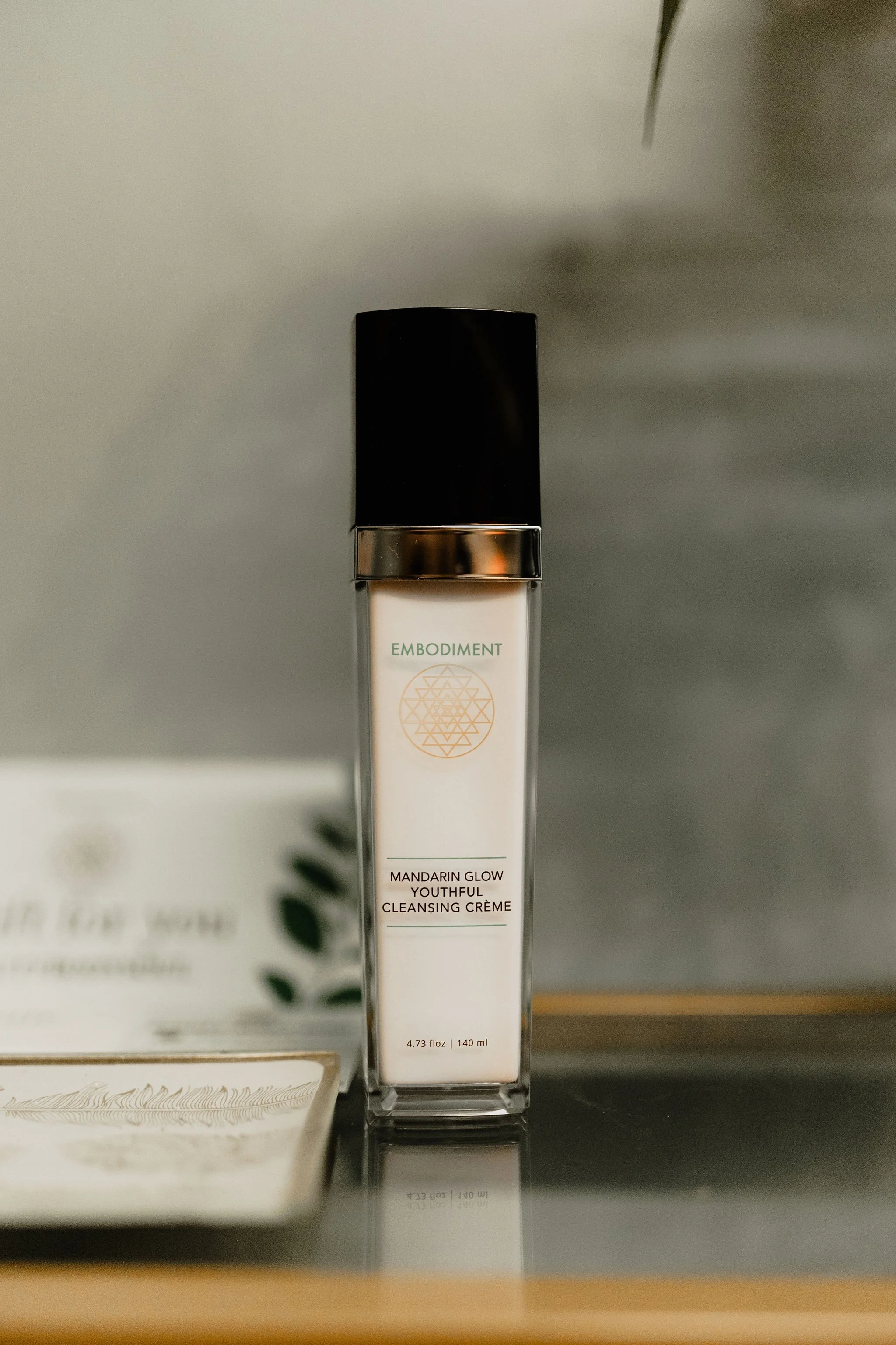 A tall, rectangular bottle of Embodiment Mandarin Glow Youthful Cleansing Crème on a reflective surface, with a blurred background.