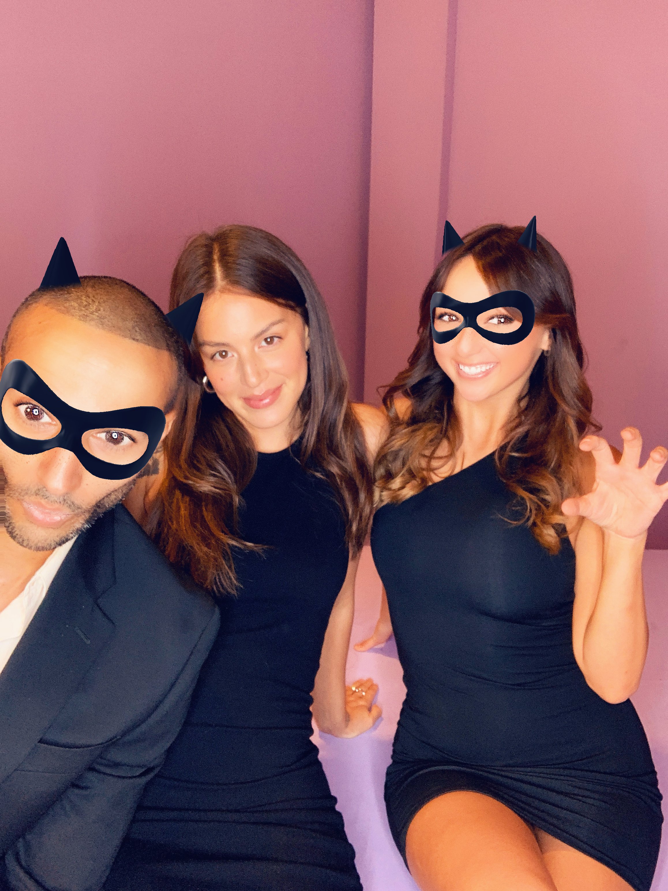 Three people wearing black outfits and cat ear masks, posing for a photo in front of a pink background, with one person making a claw gesture.