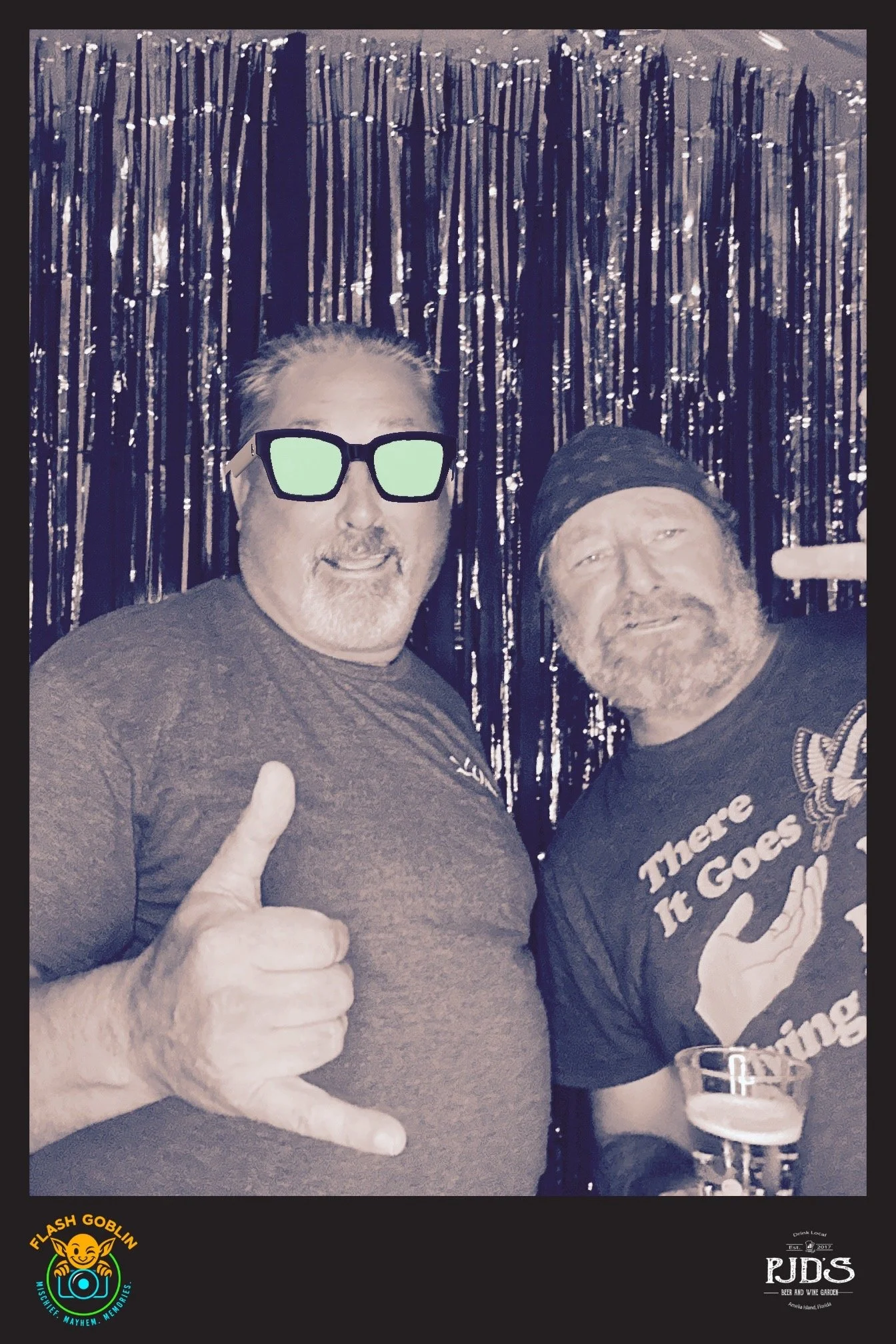 Two men smiling and posing for a photo in front of a shiny black backdrop. The man on the left is wearing sunglasses, a gray t-shirt, and giving a thumbs-up. The man on the right is wearing a black bandana and a black t-shirt with the text 'There It Goes' and a graphic of a hand holding a butterfly. The man on the right is holding a glass of beer. The bottom left corner has a logo with a smiling goblin face and the text 'Flash Goblin', and the bottom right corner has a logo with the text 'PJDS Beer and Wine Garden'.