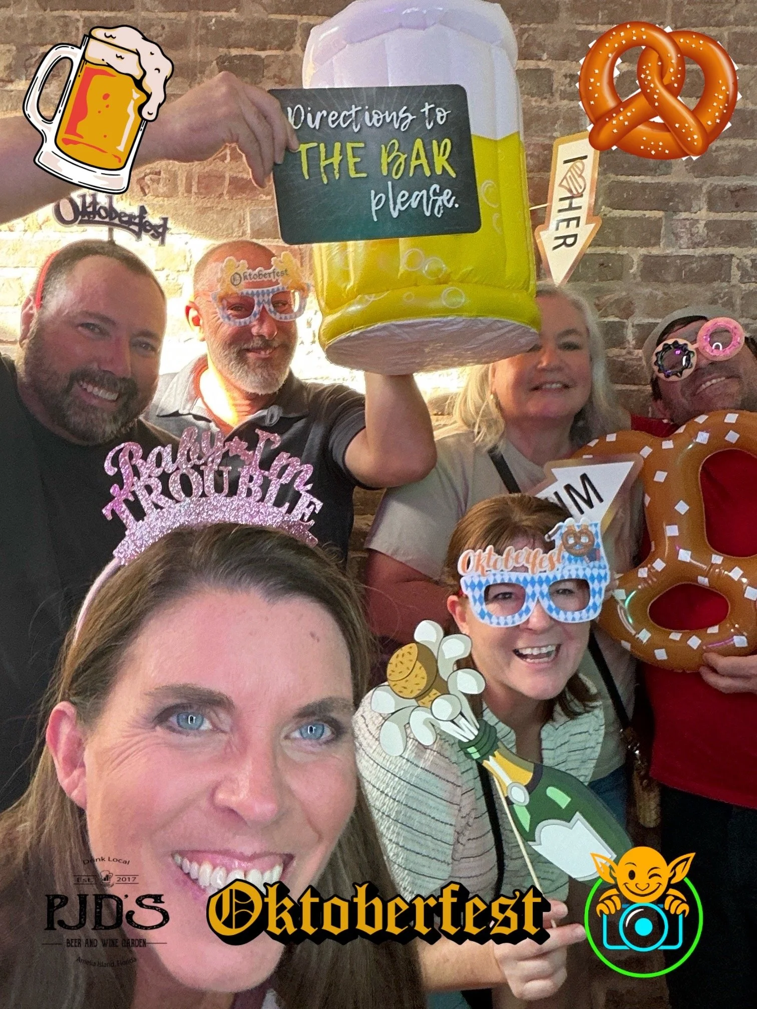 Group of people celebrating Oktoberfest indoors, with festive accessories like glasses and hats, holding a sign, a beer mug, and a pretzel, against a brick wall background.