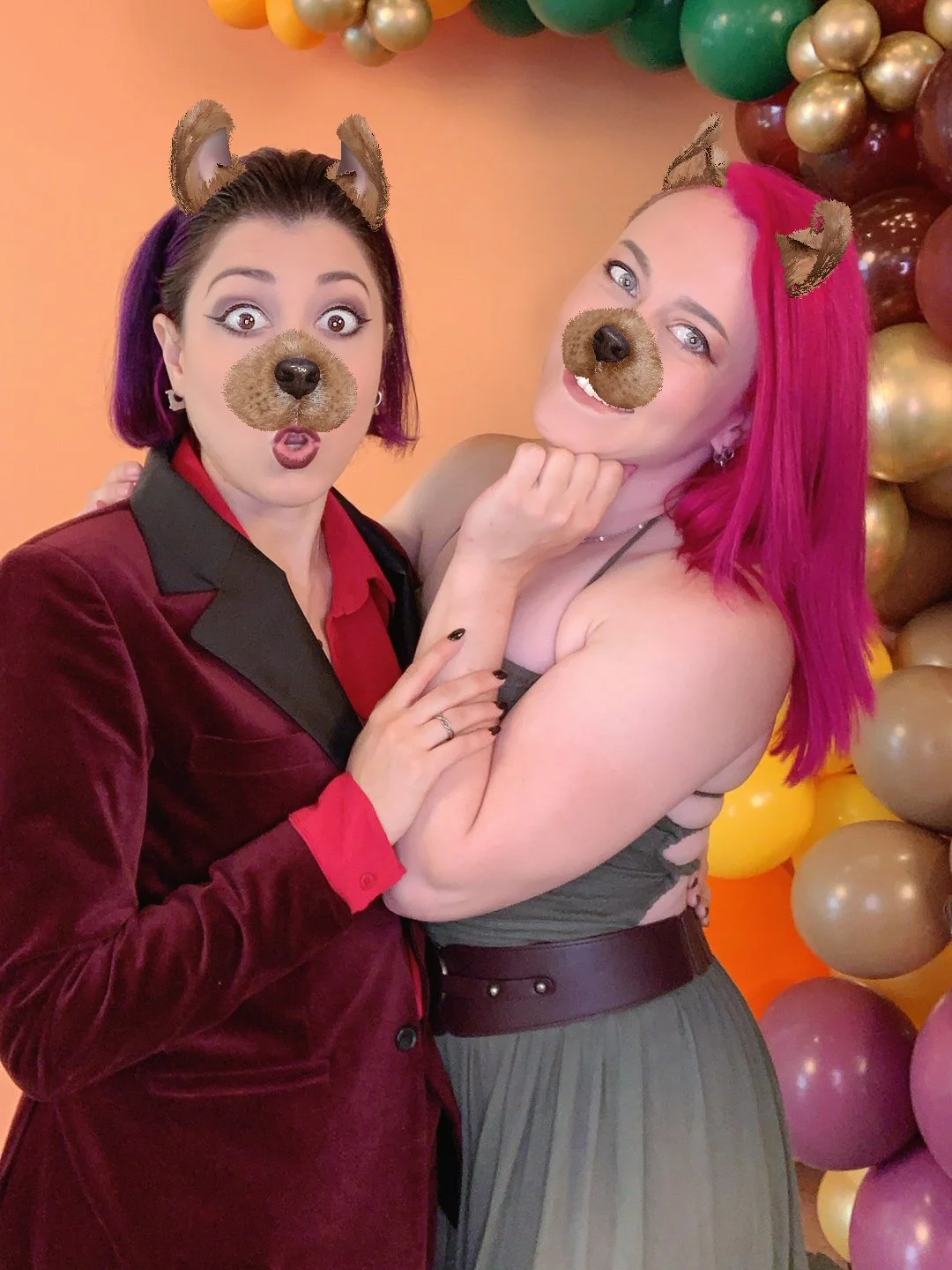 Two women with pink and purple hair using a dog face filter with ears and nose, standing in front of colorful balloons and a peach-colored background.