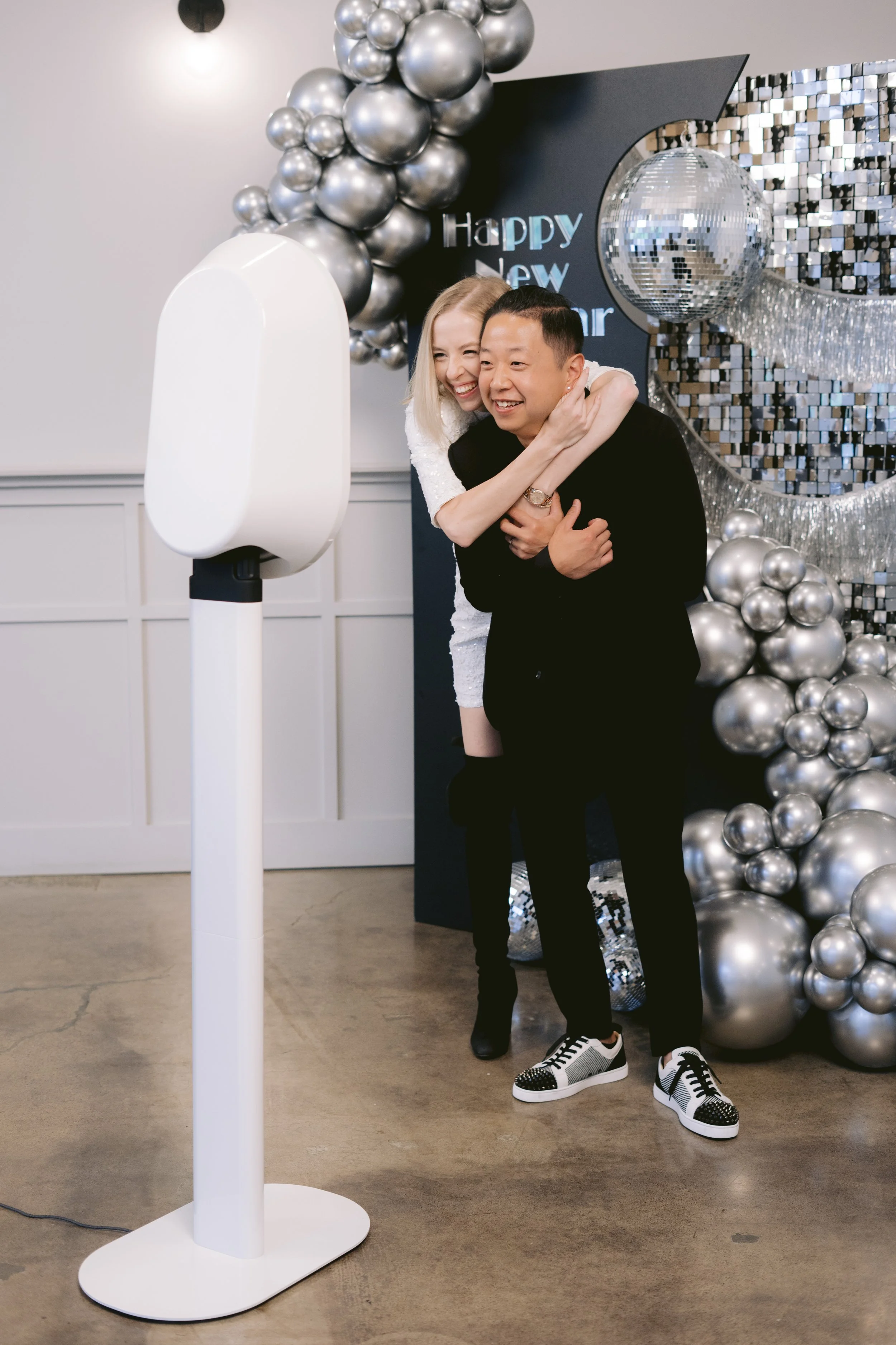 A couple taking a photo with a photo booth, smiling and hugging in front of a silver and black New Year's Eve decorations, including balloons and a shiny disco ball, with a sign that says 'Happy New Year'.