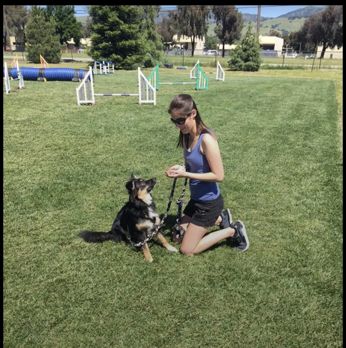 A woman kneeling on grass with a dog on a leash, in an outdoor park with agility equipment in the background.