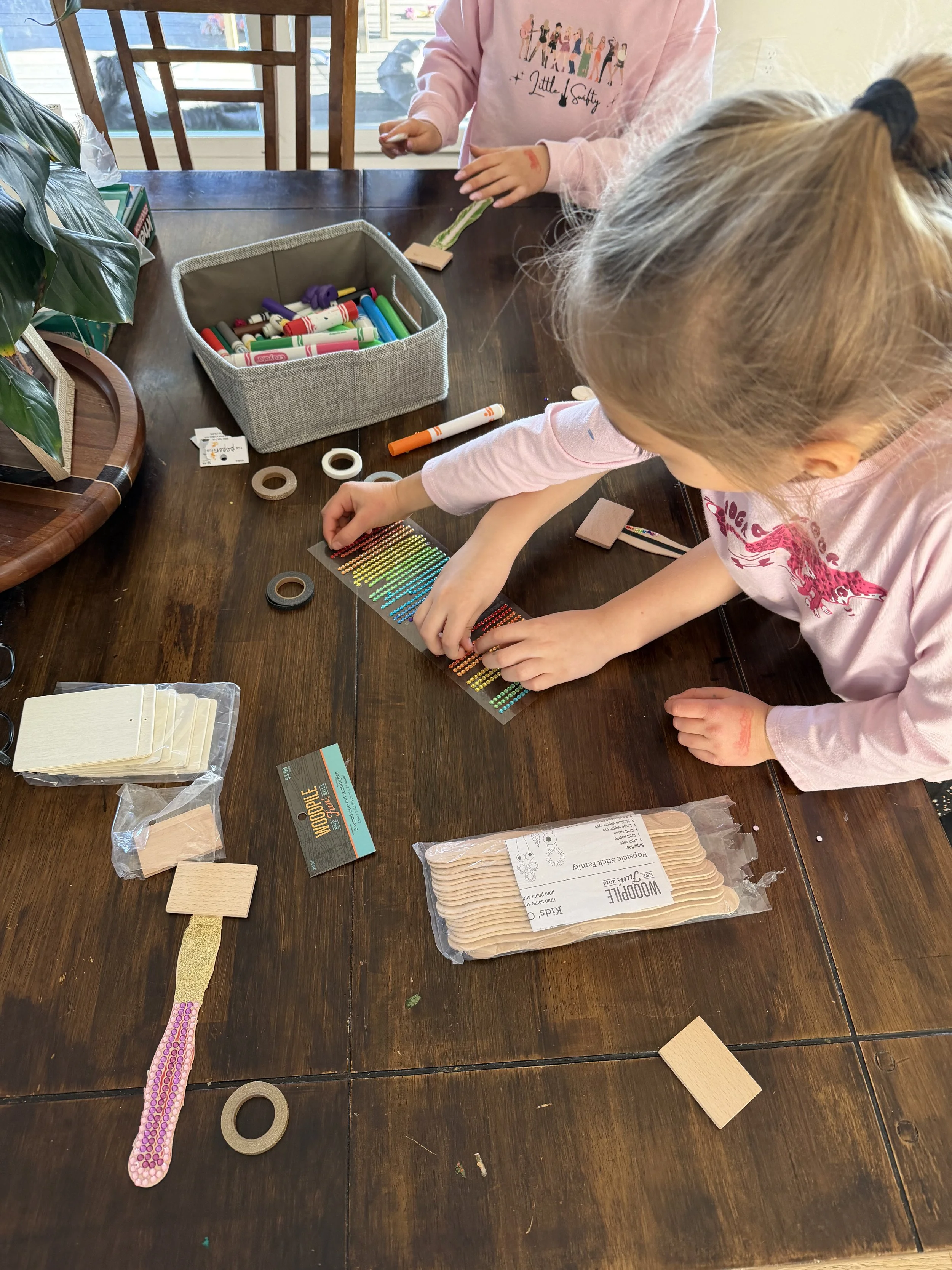 Two young children working on a craft project with colorful adhesive dots on a wooden table, surrounded by craft supplies including wooden sticks, tape, markers, and decorative tape.