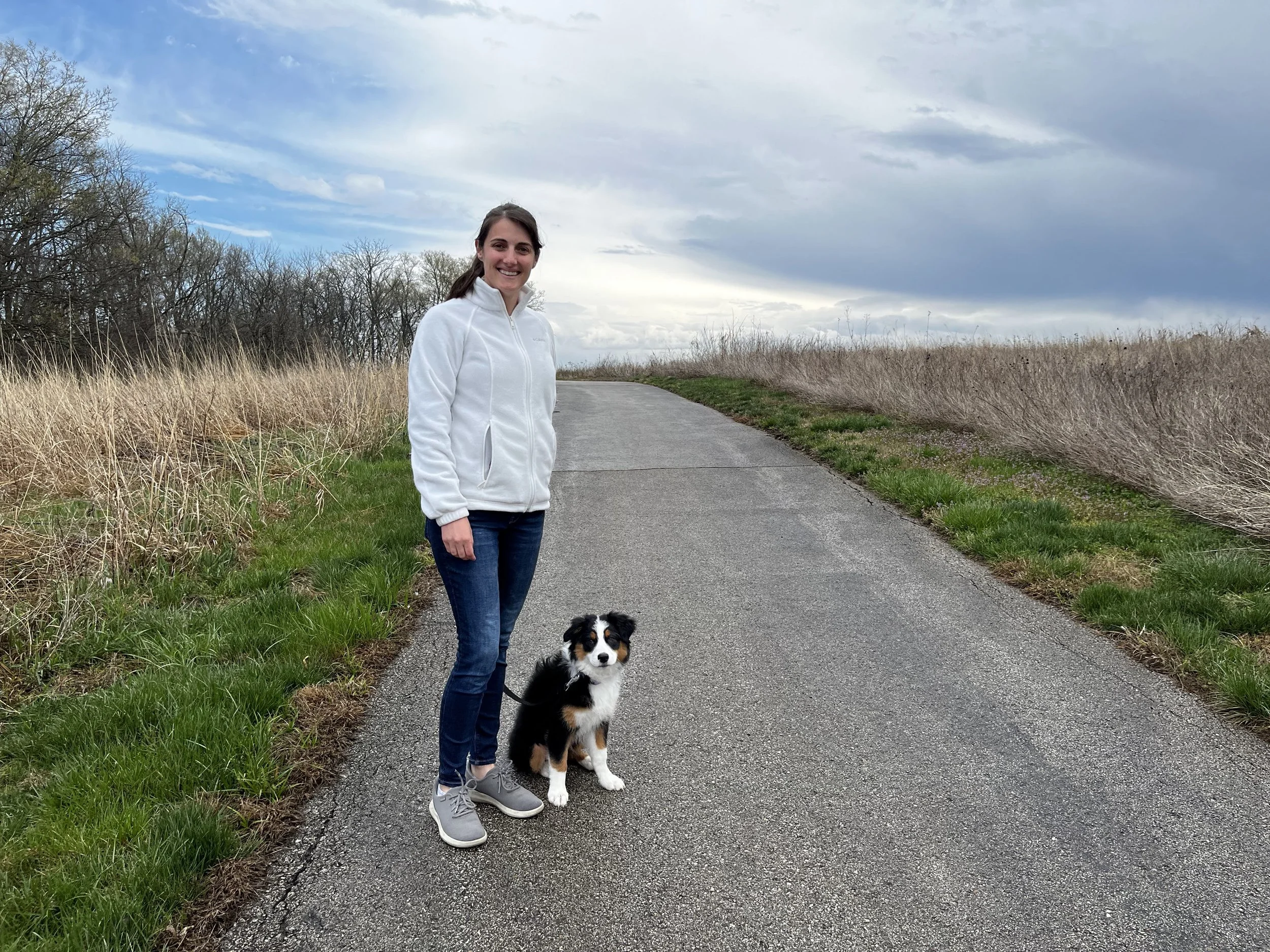 A woman in a white jacket and jeans standing on a paved path with an Australian Shepherd puppy sitting beside her in a natural landscape with tall dry grass and a cloudy sky.