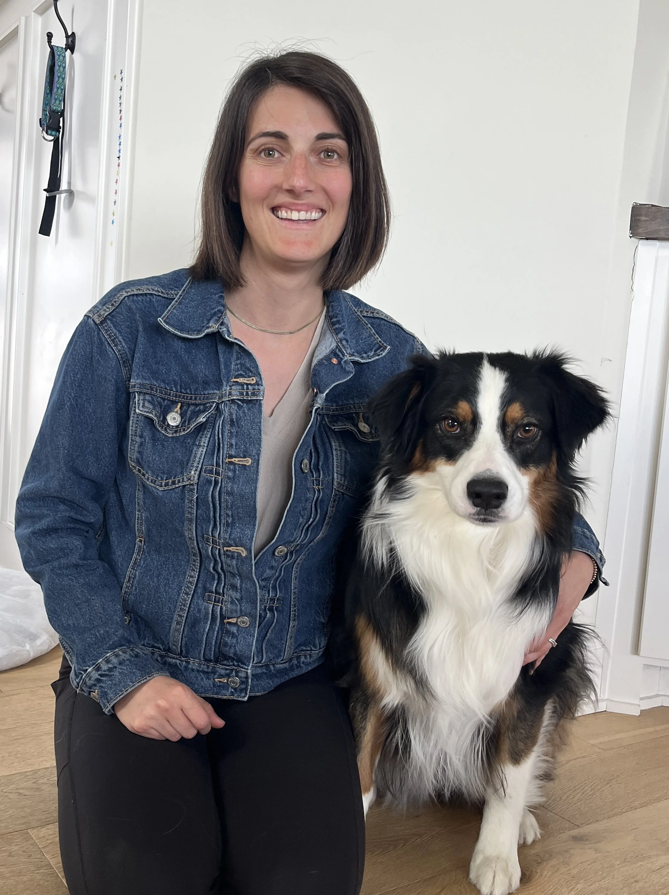 A woman with shoulder-length dark hair, wearing a denim jacket and black pants, kneels beside a large Australian Shepherd dog with black, white, and brown fur, inside a room with wooden floors and white walls.