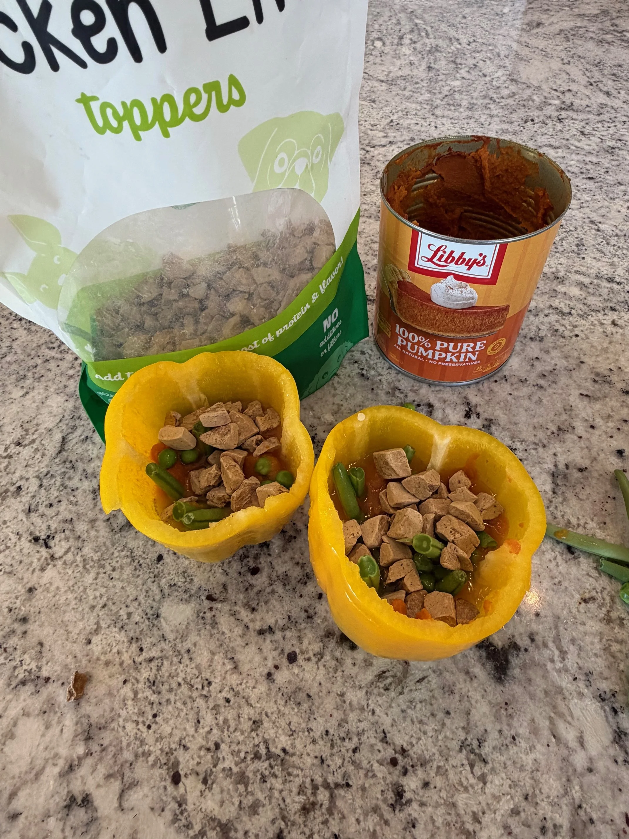 Yellow bell peppers stuffed with green beans, peas, and chopped turkey or chicken, topped with crushed crackers, on a speckled gray kitchen countertop. Behind the peppers are a bag of chicken toppers, and a can of pure pumpkin.