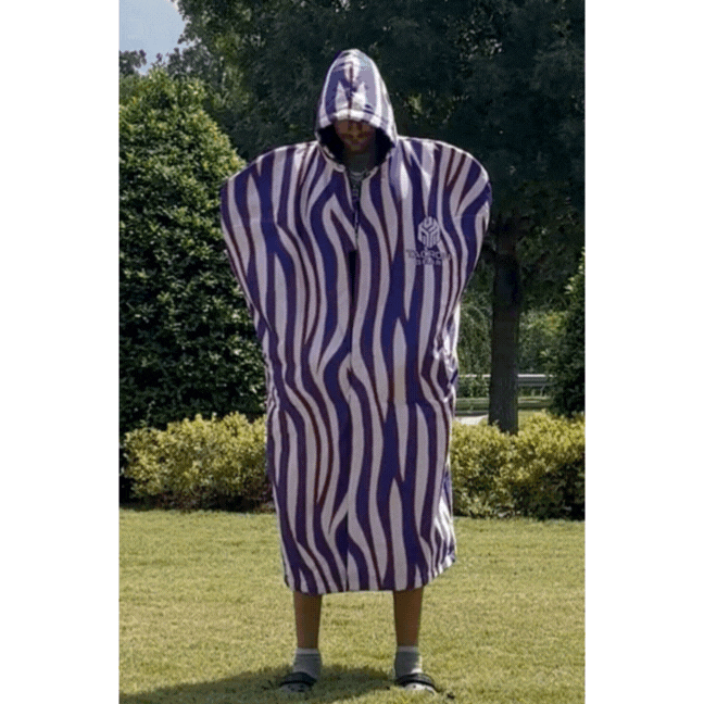 Person standing outdoors wearing a blue and white striped hooded towel with a logo, covering their head and body, on a grassy area with trees and bushes in the background.