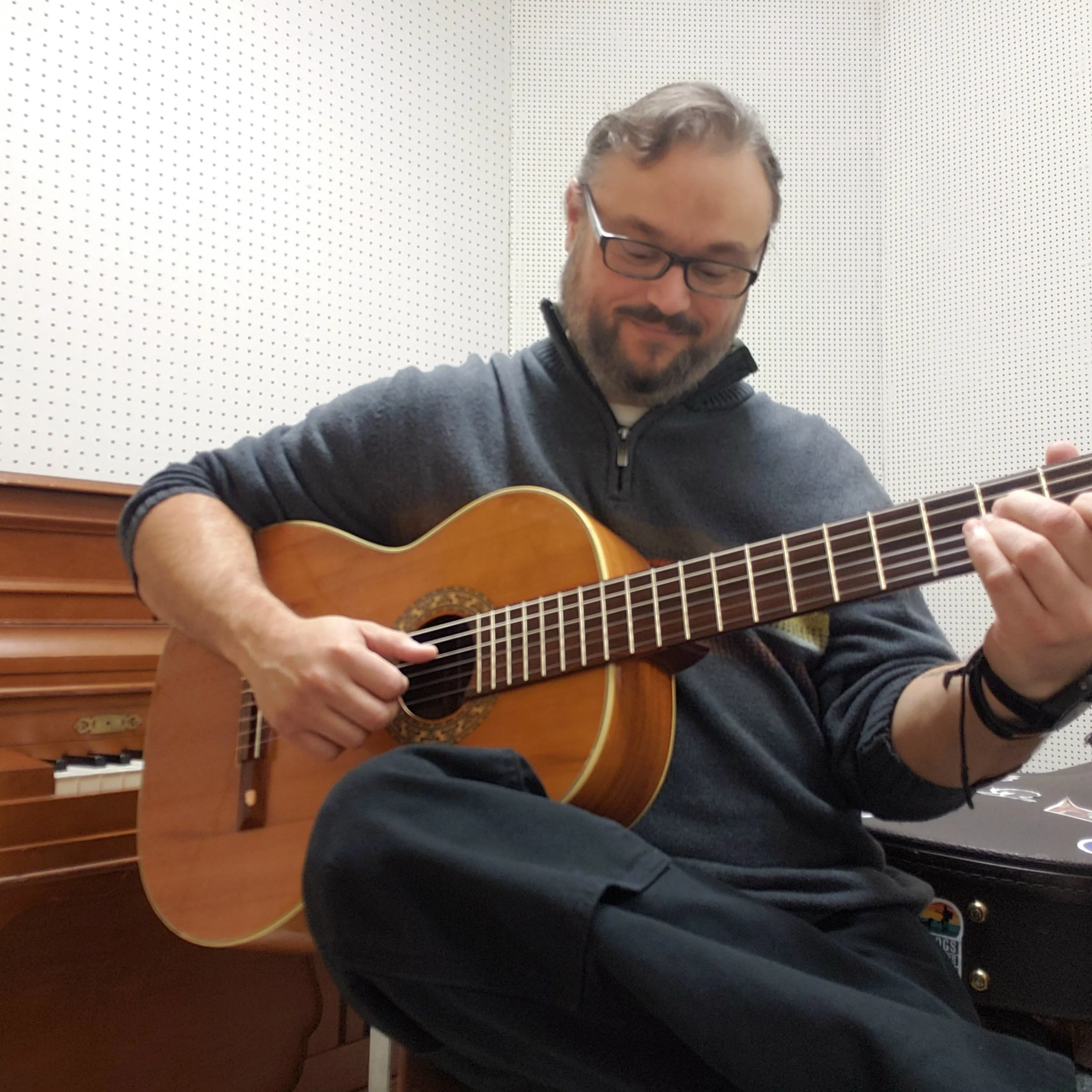 A man with glasses and a beard playing an acoustic guitar in a music studio, sitting cross-legged and smiling.