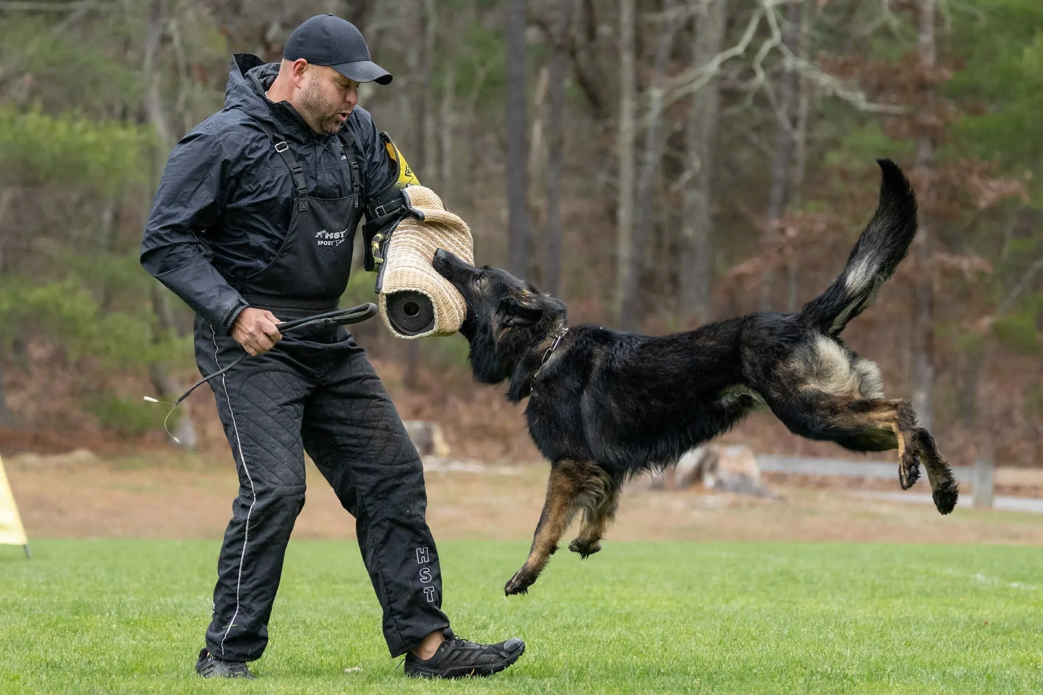 A man dressed in black training gear engaged in a dog training exercise with a German Shepherd, who is jumping up towards a protective sleeve worn by the man in an outdoor grassy area with trees in the background.