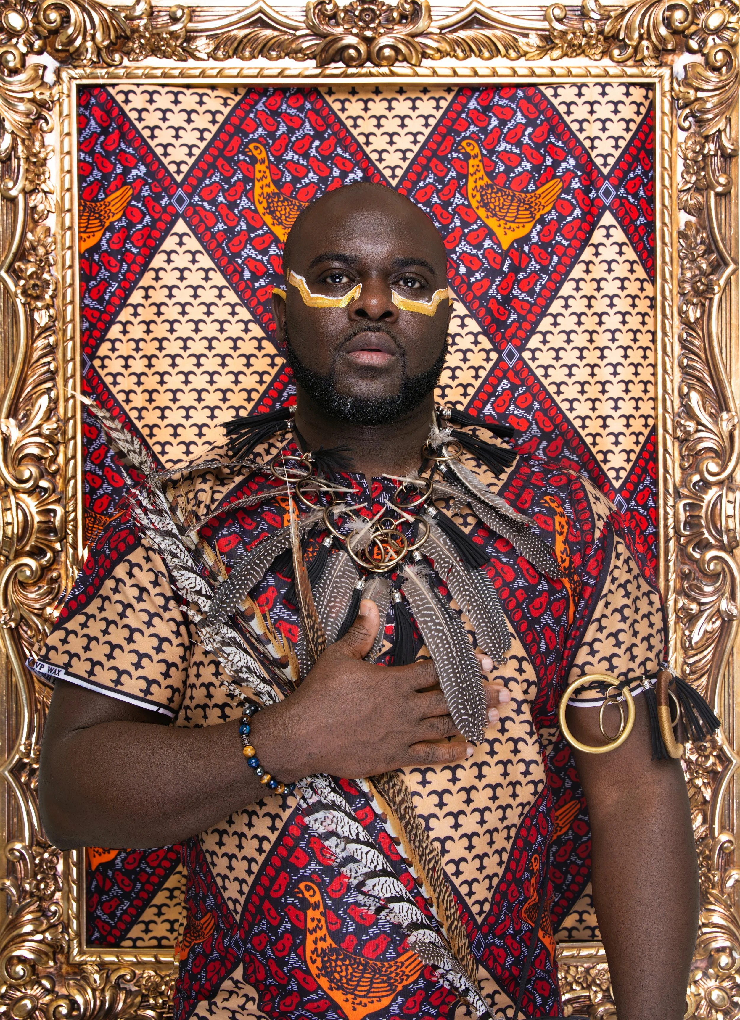 Black man with beard posing with hand on chest, wearing African-inspired jewelry and patterned attire, framed in gold with a vibrant red fabric backdrop. Born Rich: Black as My Soul portrait photography.