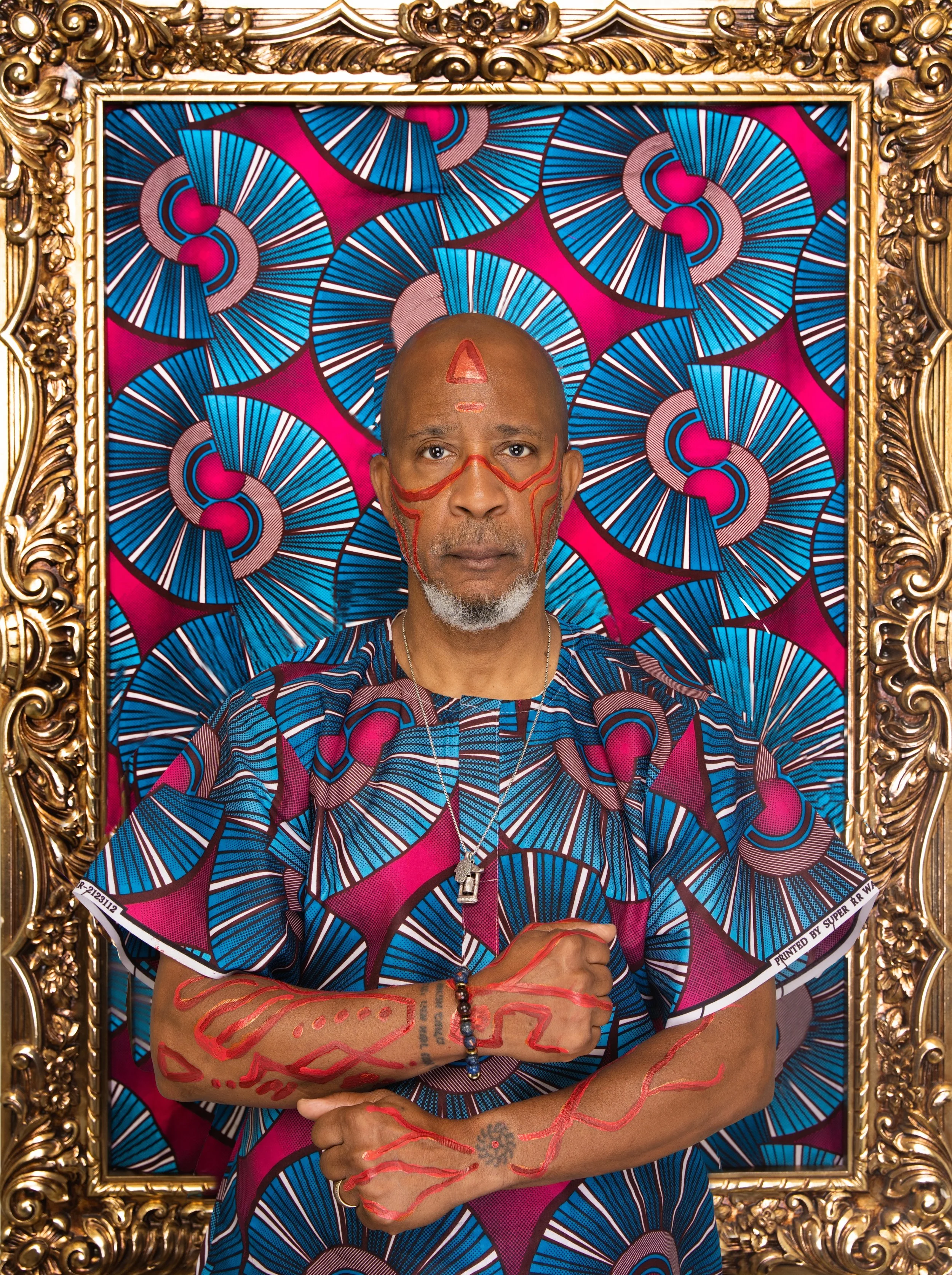 Black man with red artistic face and body paint, wearing traditional patterned African attire, posing inside an ornate gold frame with a vibrant geometric blue and pink backdrop. Born Rich: Black as My Soul photography.