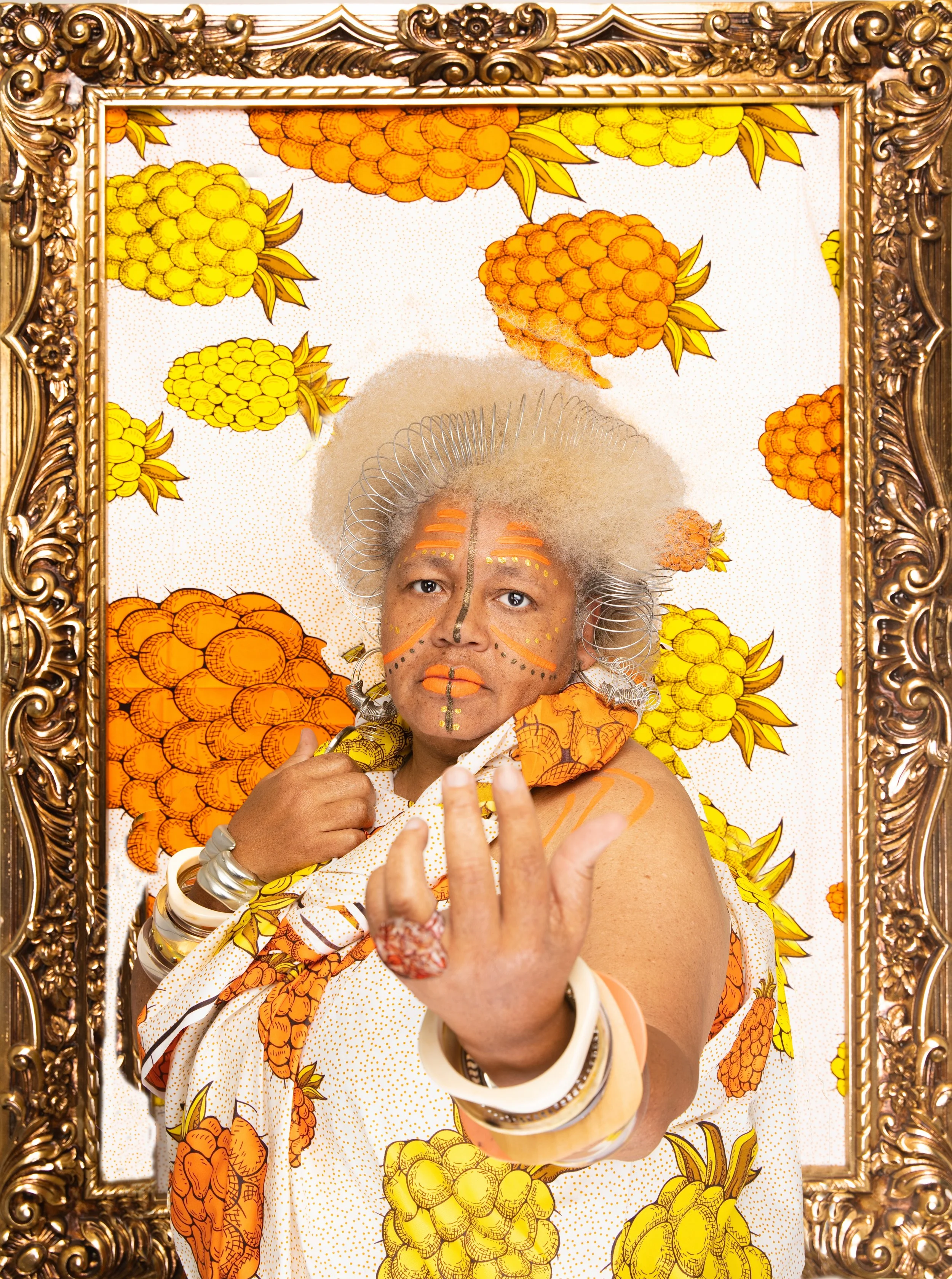 Black portrait featuring person in colorful costume and traditional white face paint, posing inside an ornate gold frame against a yellow-orange pineapple-patterned backdrop. Born Rich: Black as My Soul cultural photography.