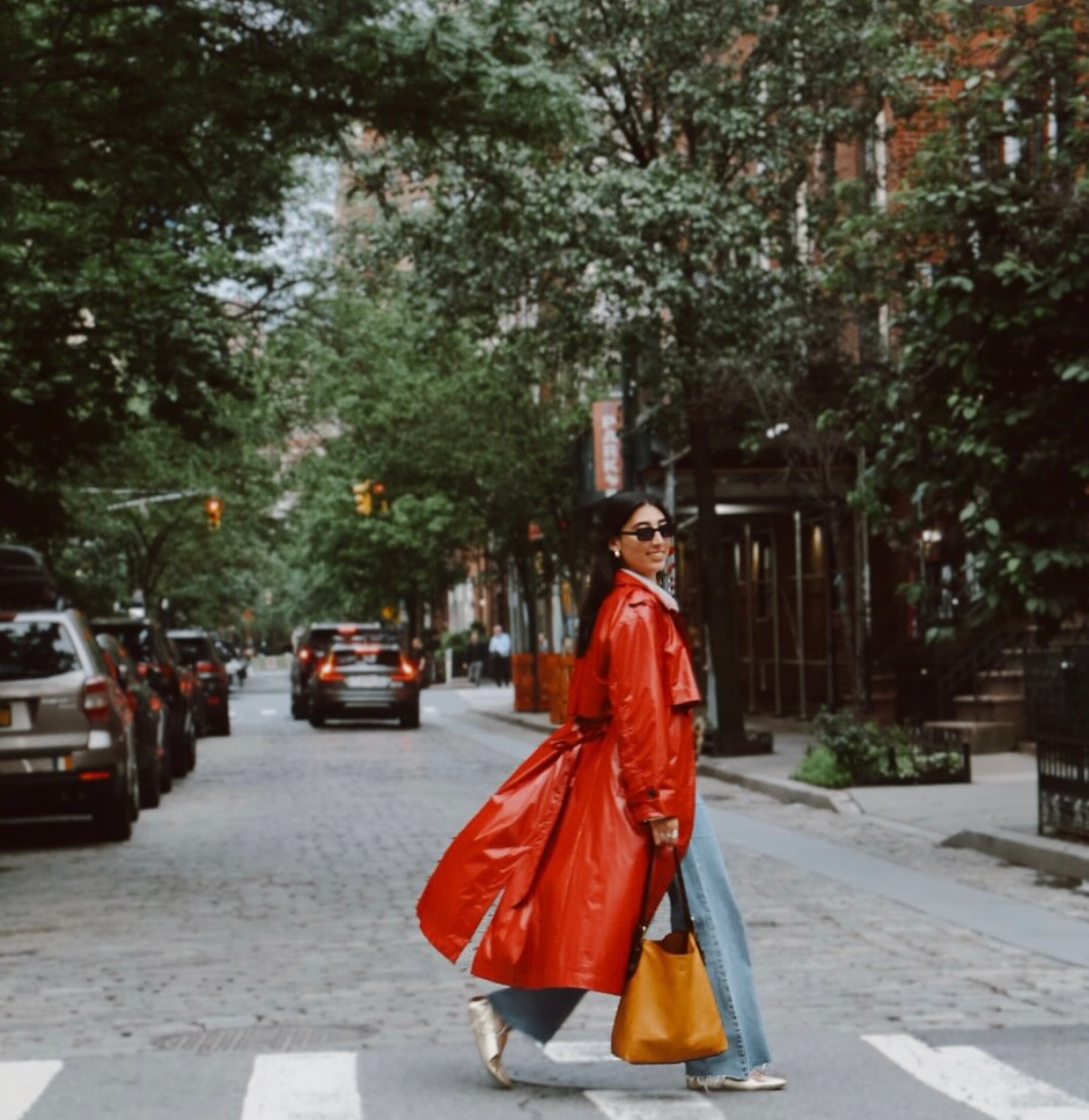 Woman crossing the street wearing a red trench coat, sunglasses, and black pants, carrying a mustard-colored bag with a city street background.