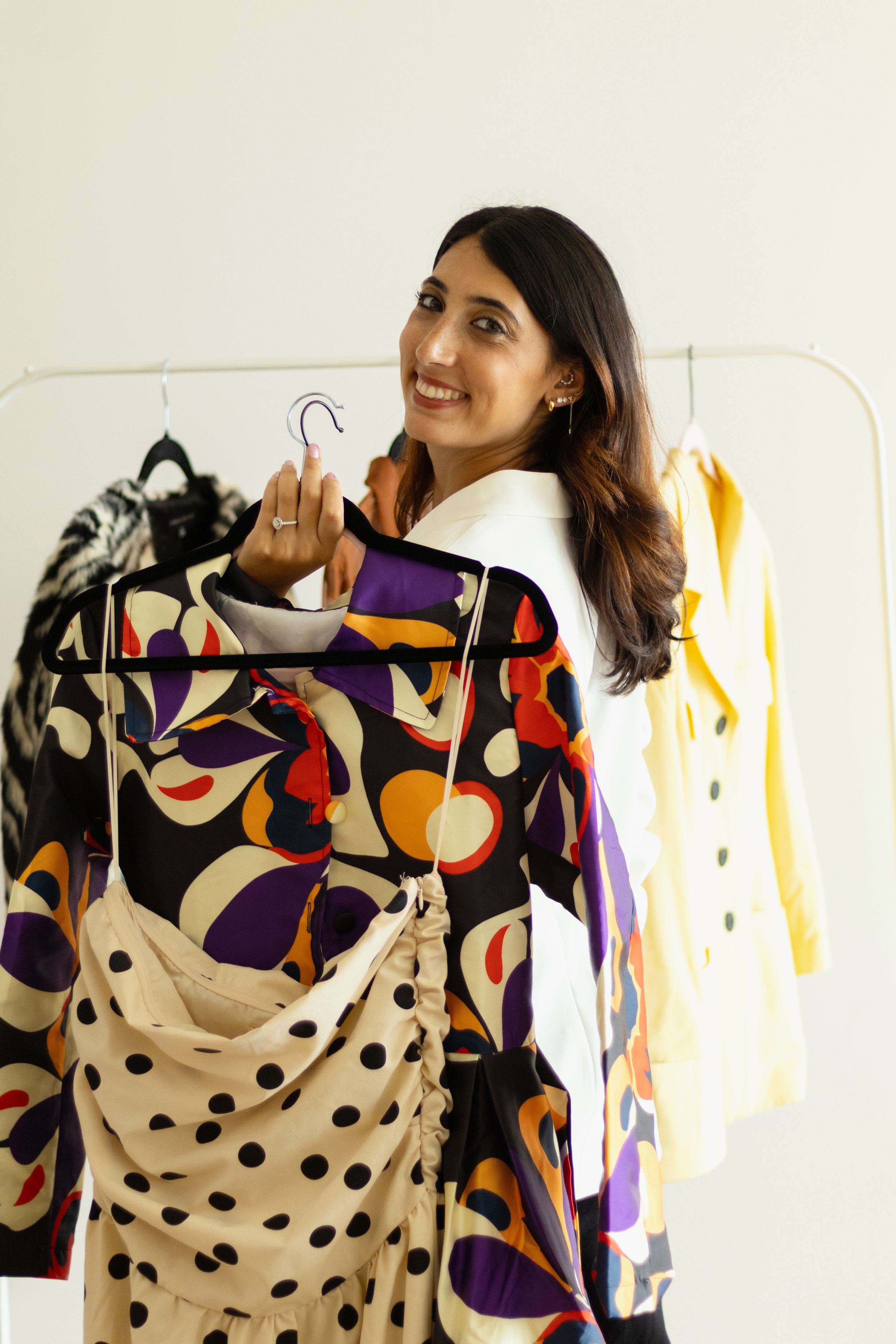 A woman smiling as she holds a black clothing hanger with a multicolored dress, standing in front of a clothing rack with various garments, including yellow and animal print pieces.