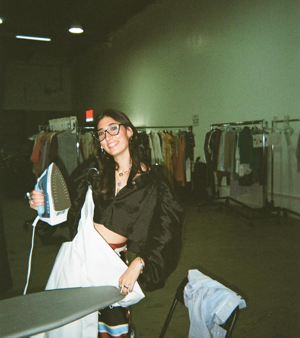 A young woman with dark hair and glasses smiling while holding an iron and a white cloth in a clothing store or warehouse with racks of clothing in the background.