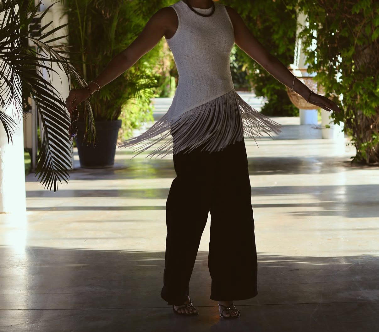 Person wearing a sleeveless white top with fringe, black wide-leg pants, and heeled sandals, standing outdoors surrounded by greenery, with arms extended to the sides.