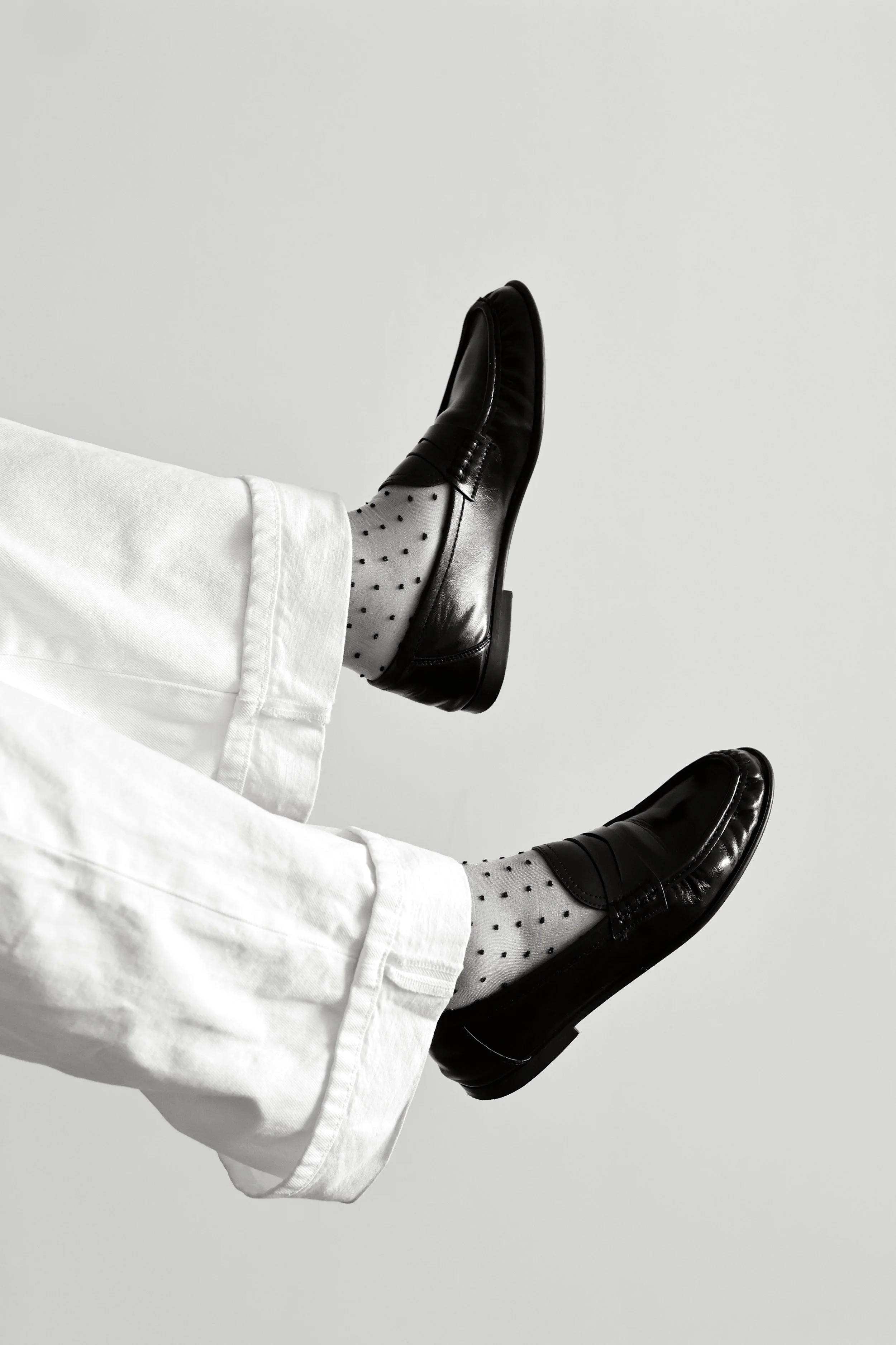 Close-up of a person's legs wearing white pants, polka dot socks, and black shiny loafers, with the person reclining on a plain white background.