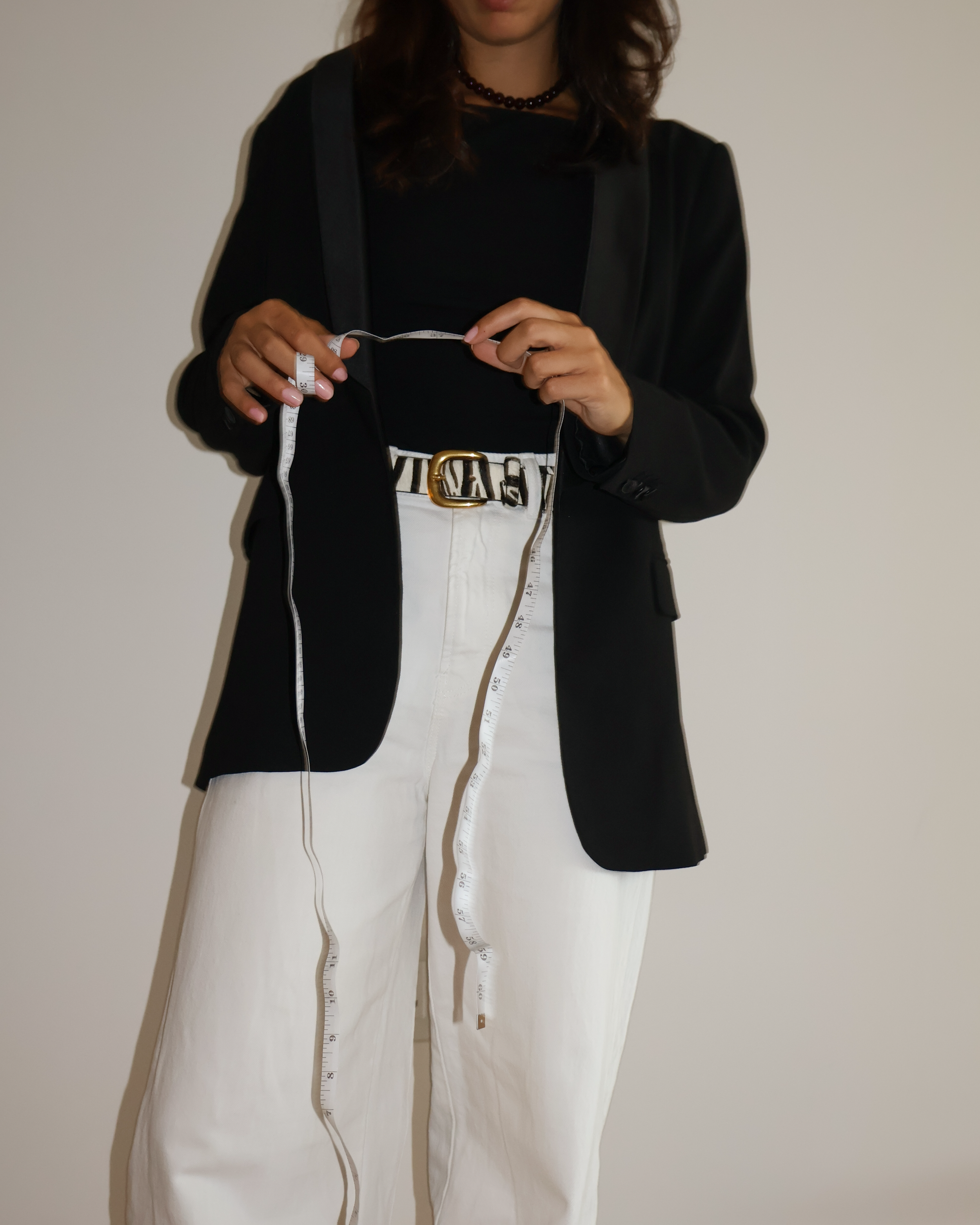 A woman in a black blazer and white pants holding a measuring tape in front of a plain wall.