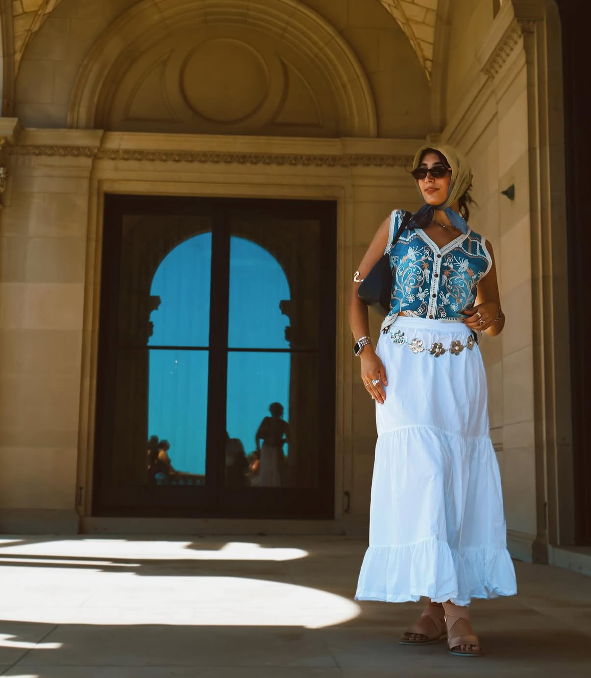 A woman wearing sunglasses, a beige headscarf, a blue embroidered sleeveless top, and a long white skirt with floral embellishments, standing in front of a historic stone building with a large arched window. There are people reflected in the window, and sunlight creates shadows on the ground.