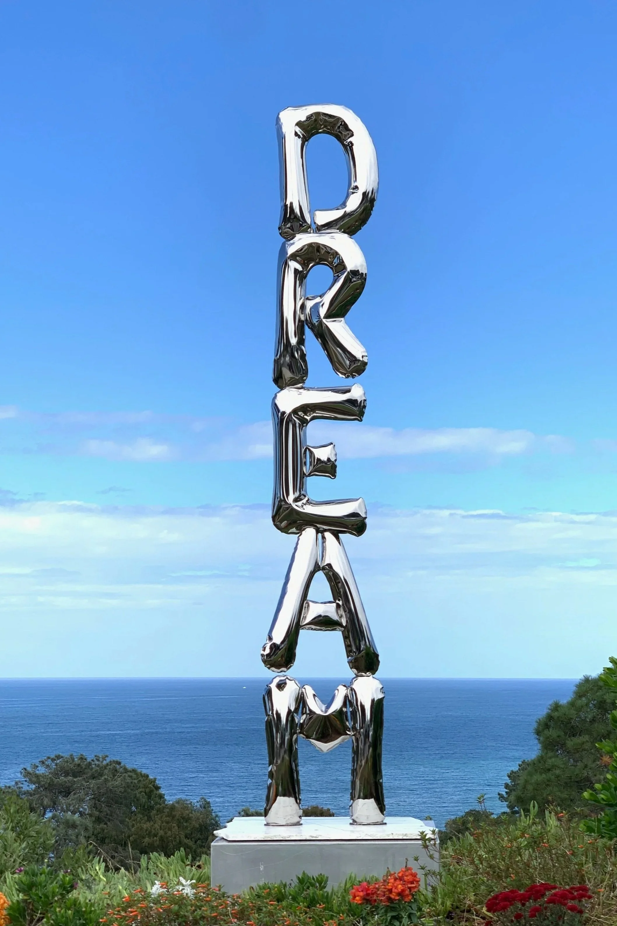 Sculpture spelling out 'DREAM' in metallic letters, with the ocean and blue sky in the background
