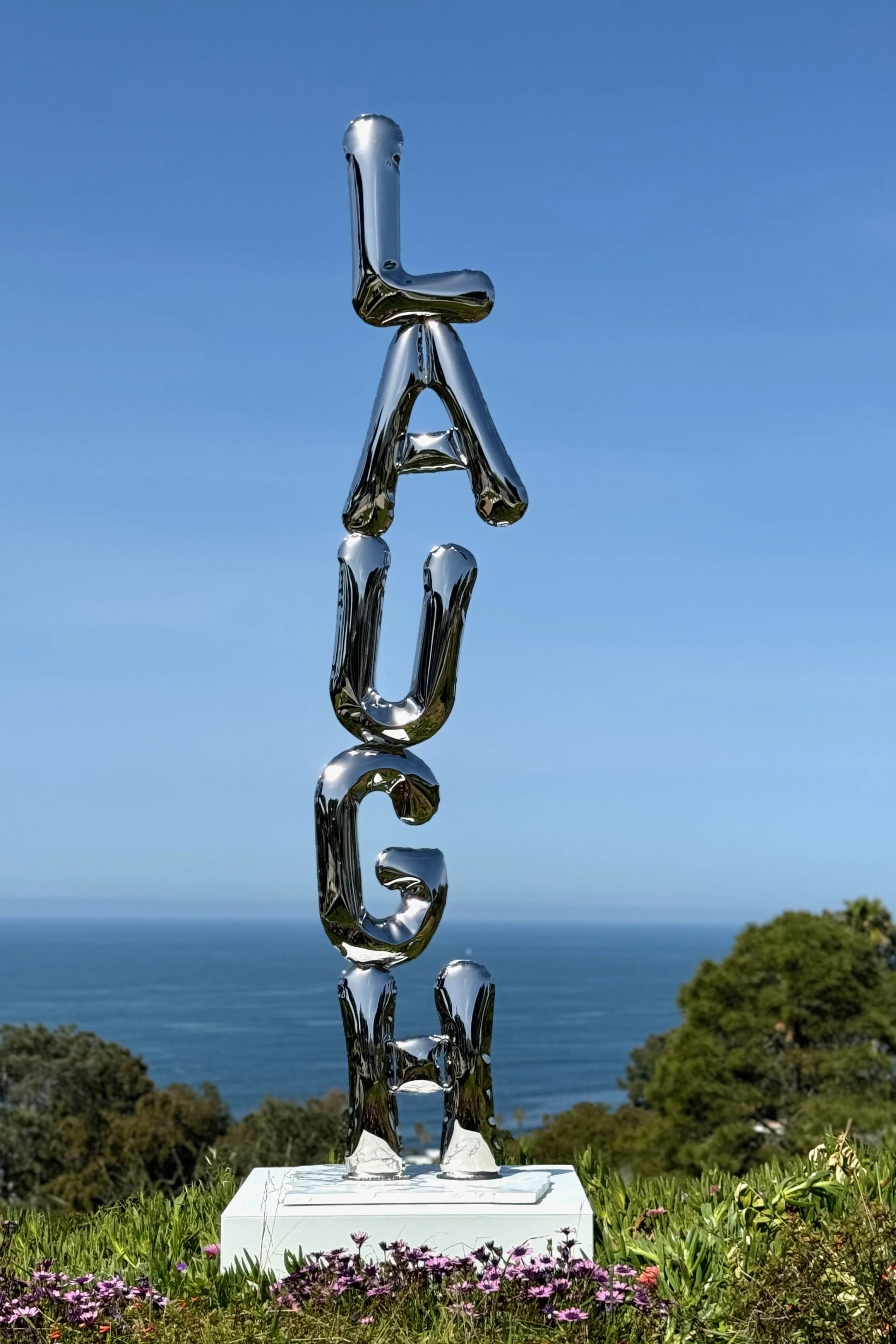 A large metallic sculpture spelling the word "Laugh" stacked vertically, set outdoors with trees, flowers, and the ocean in the background under a blue sky.