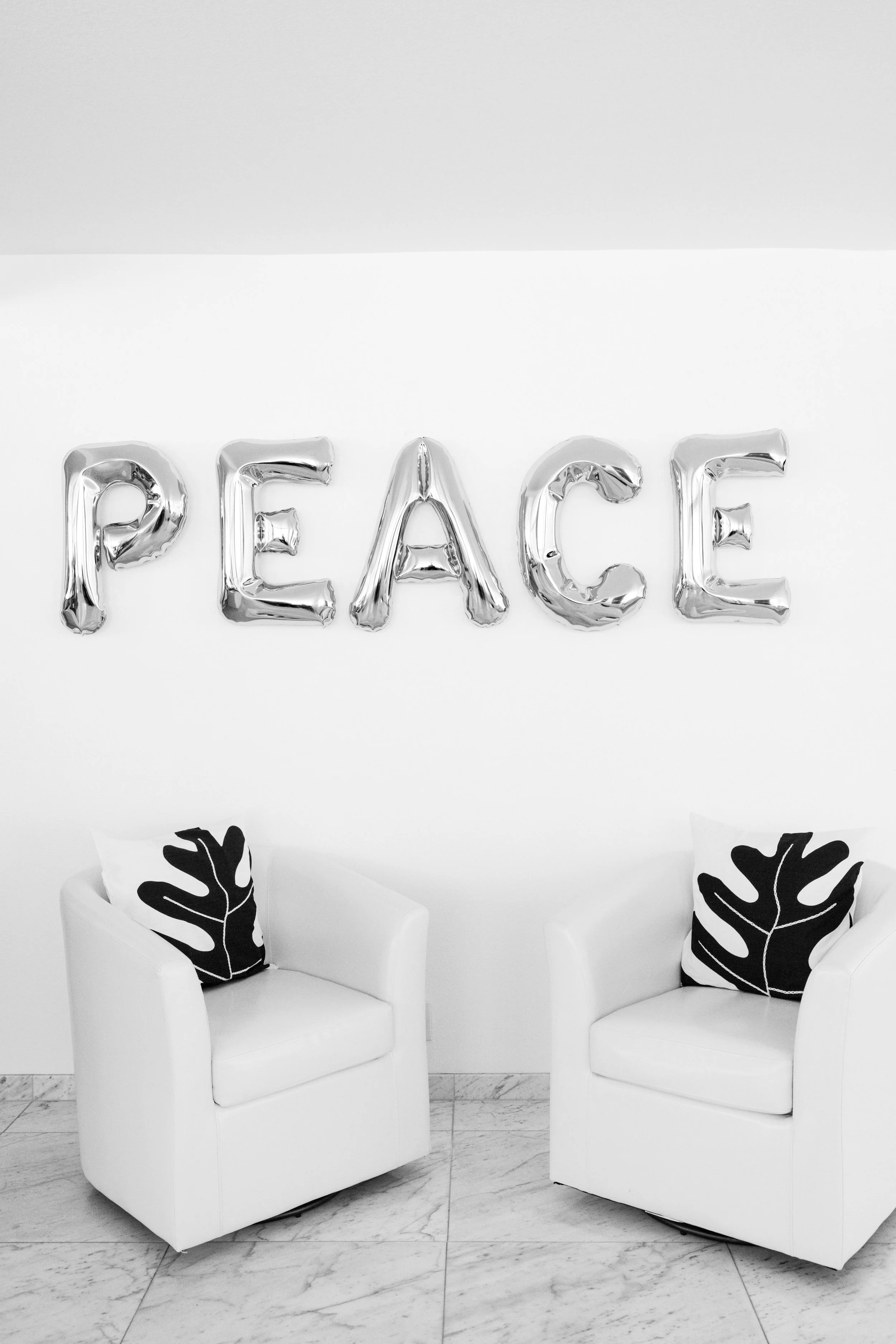 A large metallic sculpture spelling the word "PEACE" stacked vertically, set indoors with two white barrel chairs on white marble floor