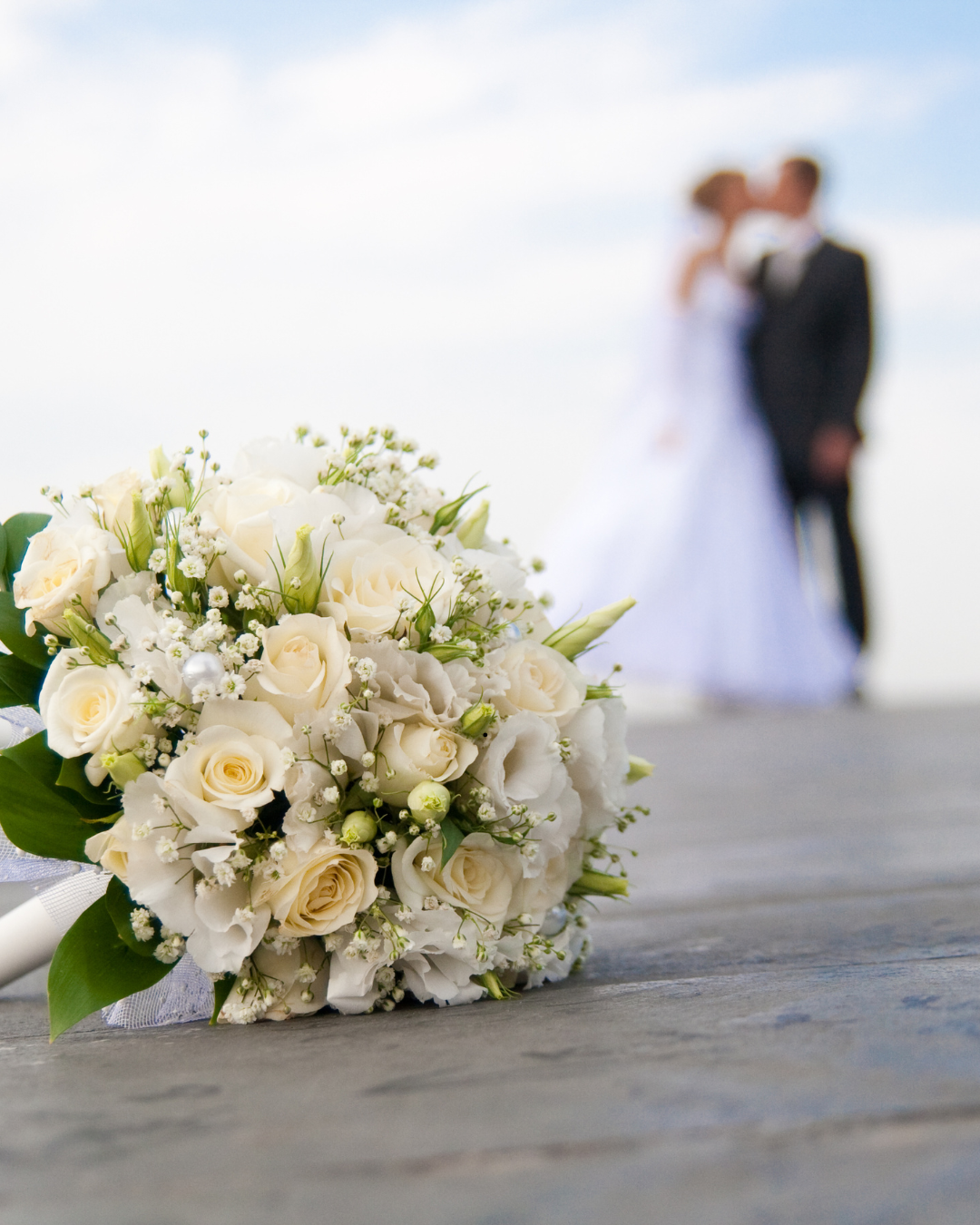 A bridal bouquet of white roses and flowers lying on a wooden surface with a blurred bride and groom kissing in the background.