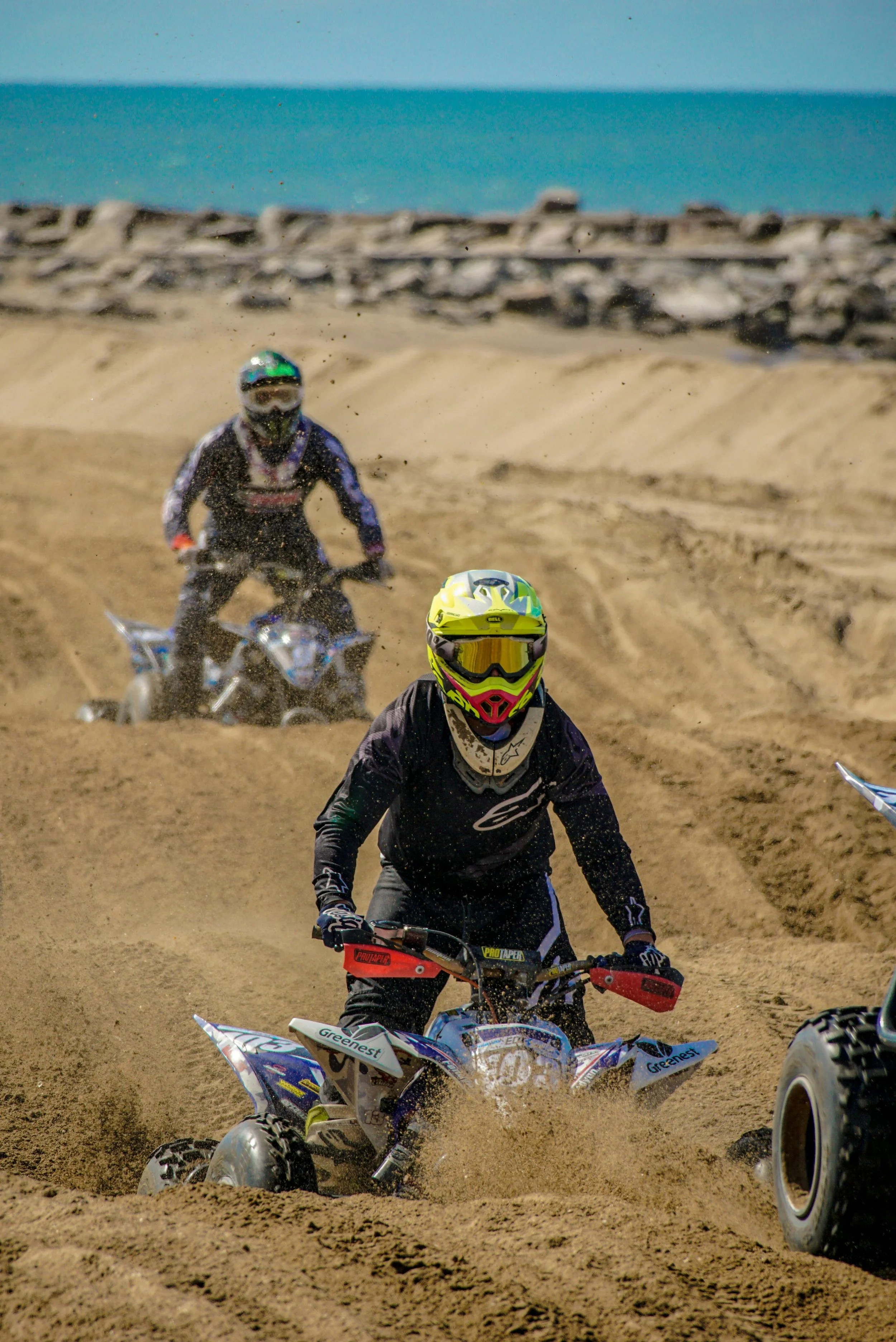 Two motocross riders riding on a sandy dirt track with the ocean in the background; the rider in the foreground is wearing a black outfit and a yellow helmet, and the rider in the background is wearing a black outfit with a helmet and goggles.