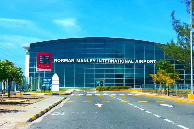 Front view of Norman Manley International Airport building with a sign, surrounded by palm trees and a clear blue sky.