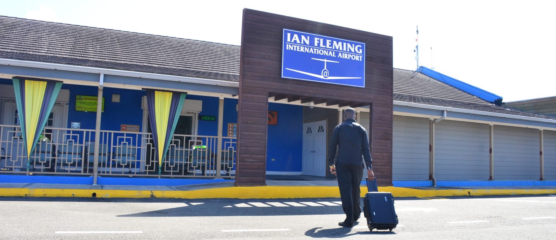 A man with a suitcase walking towards the entrance of Ian Fleming International Airport, which has a large sign displaying the airport's name and logo, with a mostly blue and yellow color scheme.