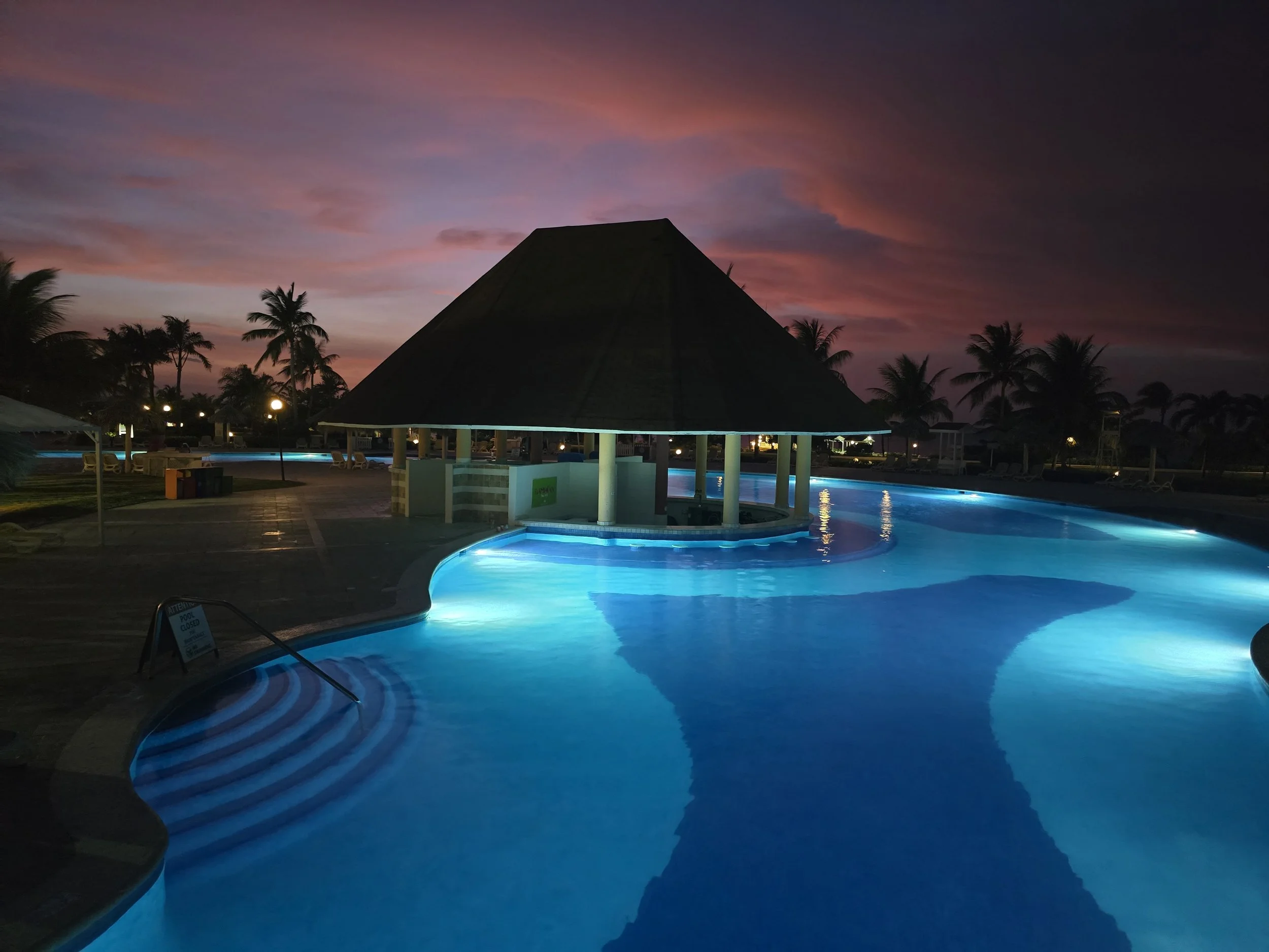 View of a lit outdoor swimming pool at sunset with a shaded structure and palm trees in the background.