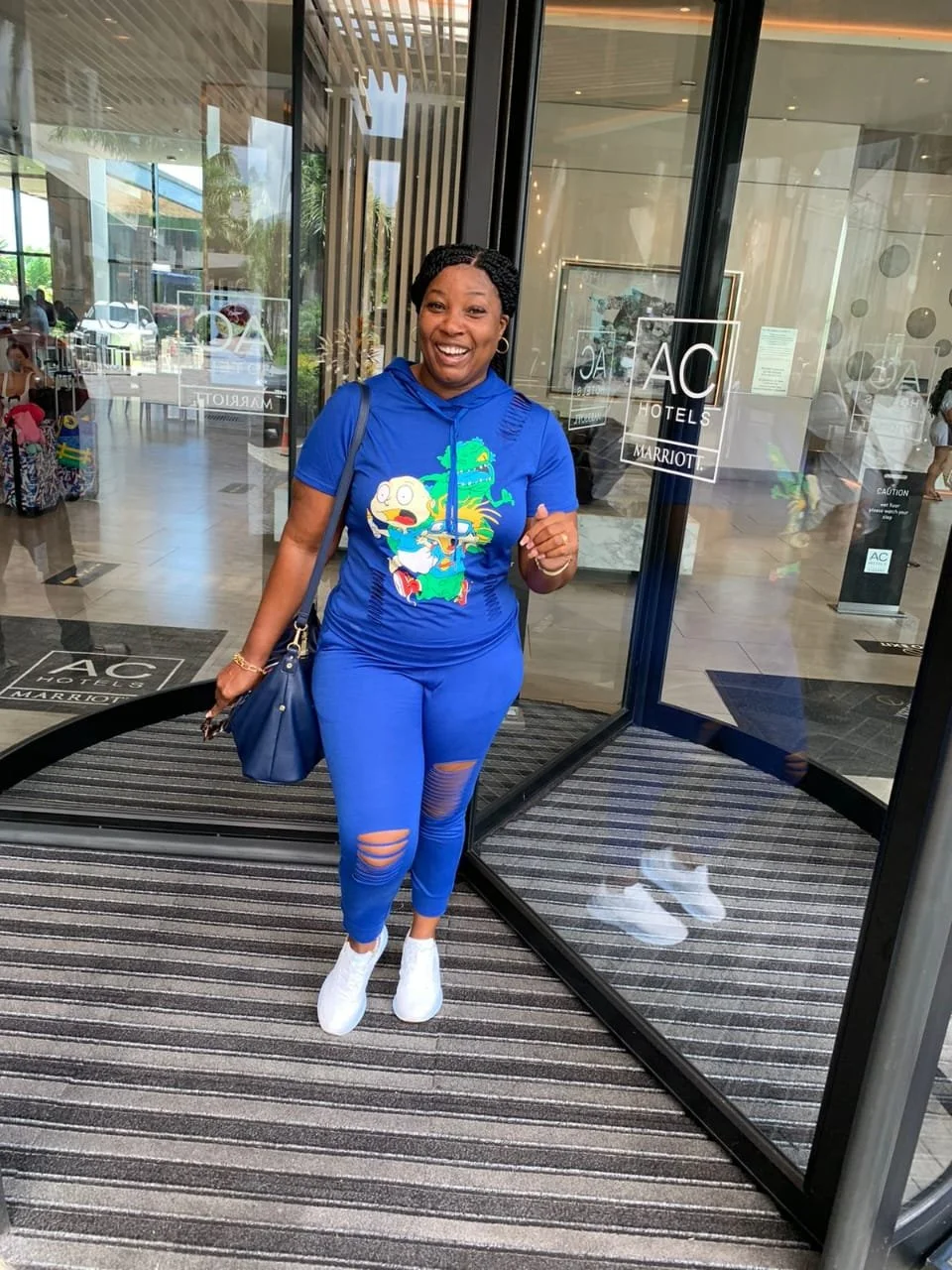 A woman in a bright blue t-shirt and matching distressed pants standing at the entrance of a hotel with glass doors, smiling, with a luggage cart and other hotel guests visible in the background.