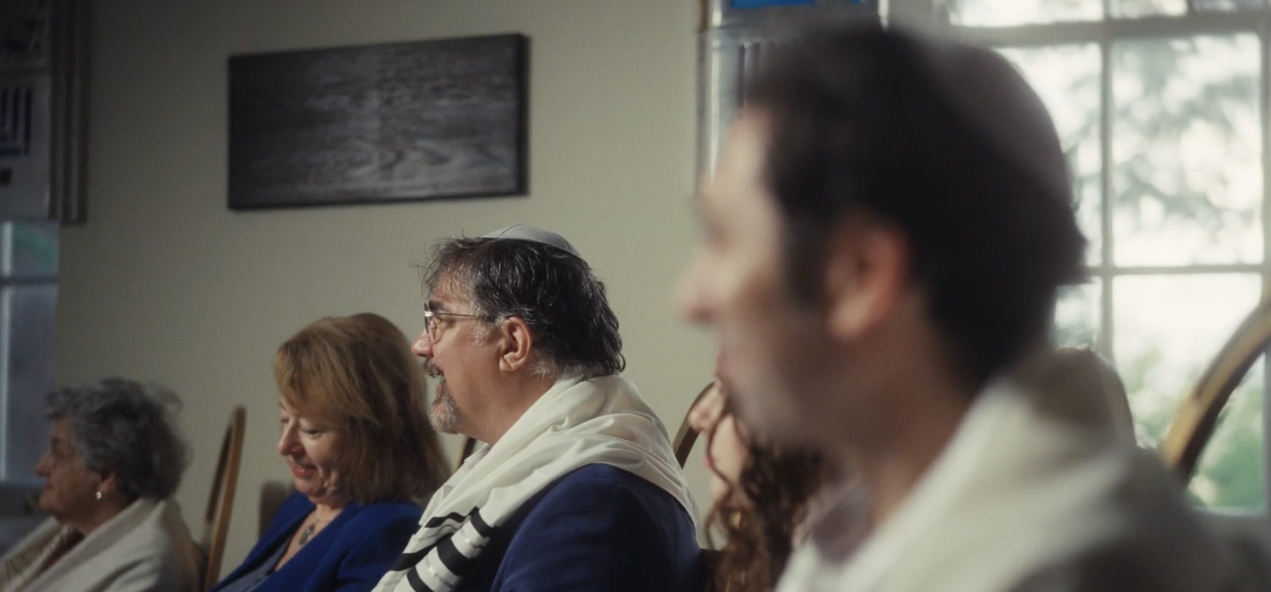 People sitting in a room during a gathering or ceremony, with natural light coming through a large window.